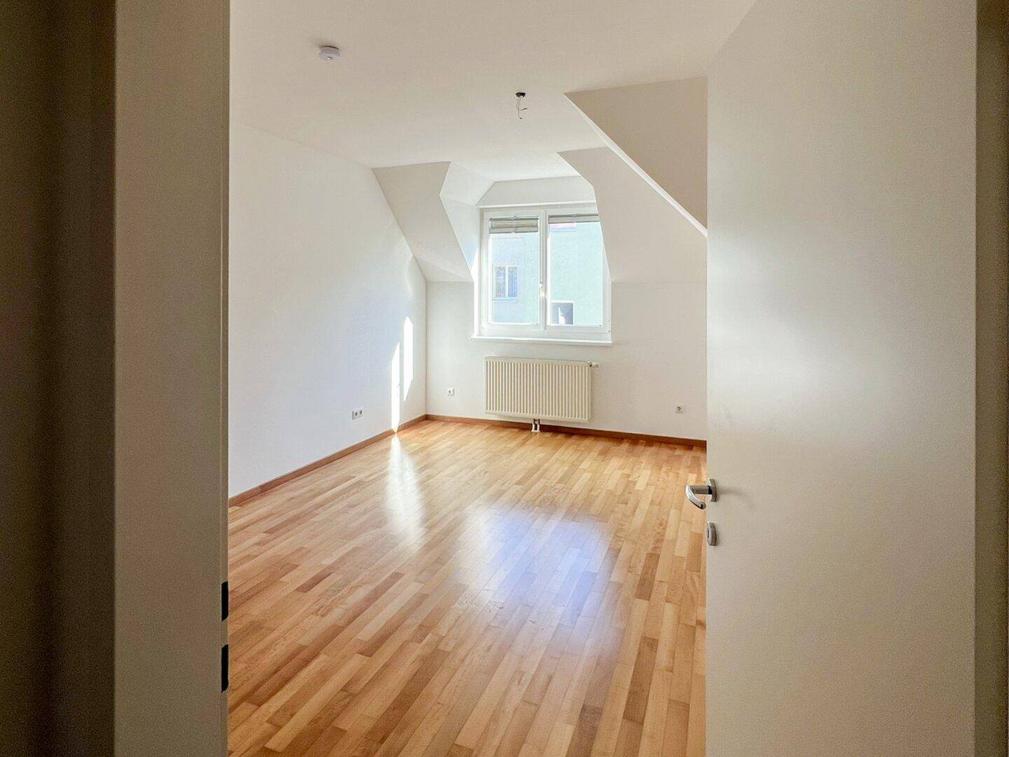 Helles Zimmer im Dachgeschoss mit schrägen Wänden, Parkettboden und einem Fenster.