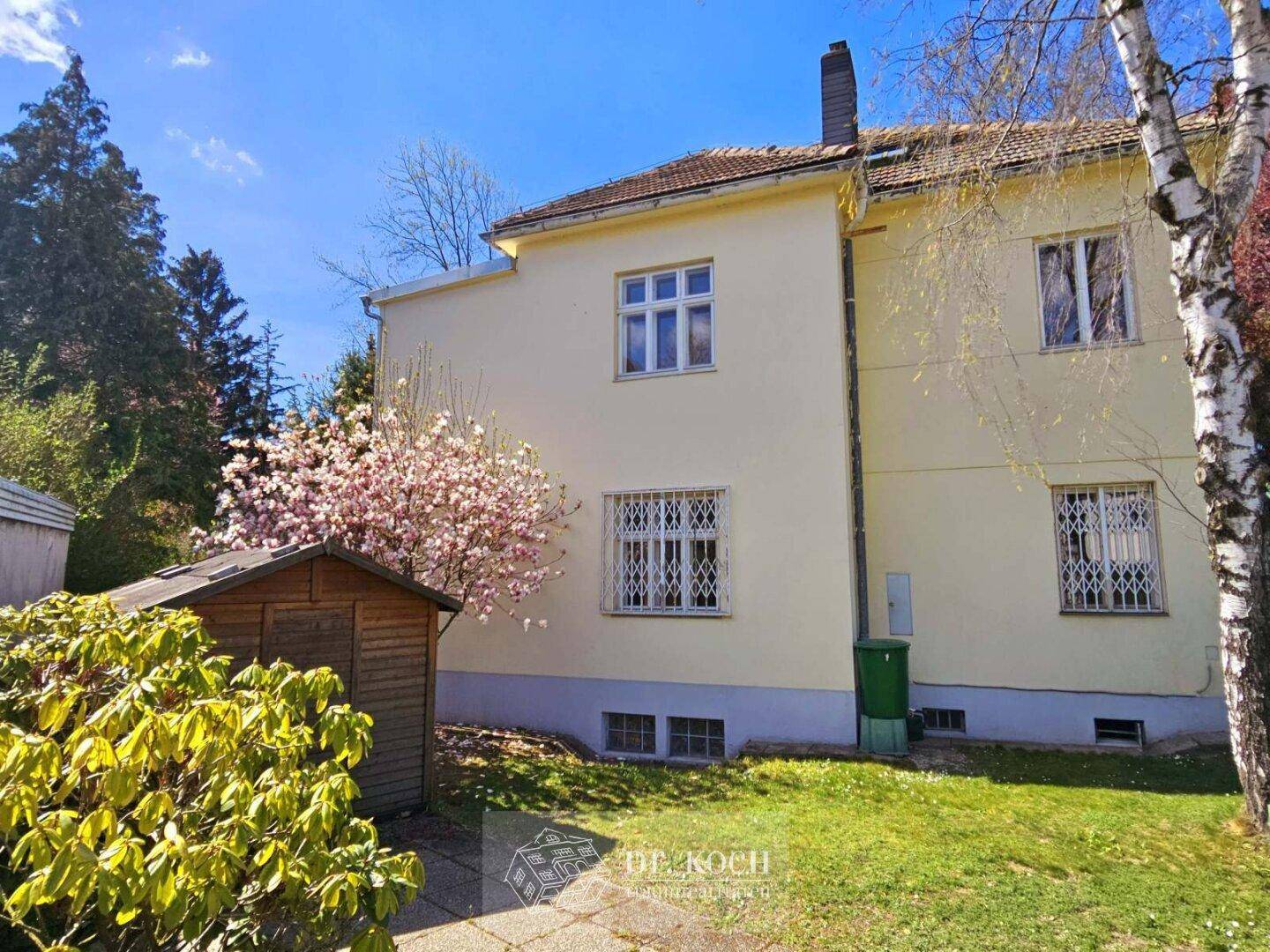 Rückansicht des Hauses mit Garten, Schuppen und blühendem Baum an einem sonnigen Tag.