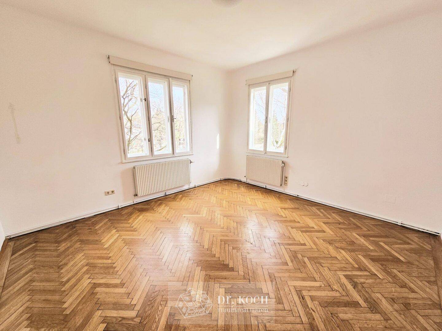 Helles Zimmer mit Fischgrätparkettboden und zwei großen Fenstern, die viel Tageslicht hereinlassen.