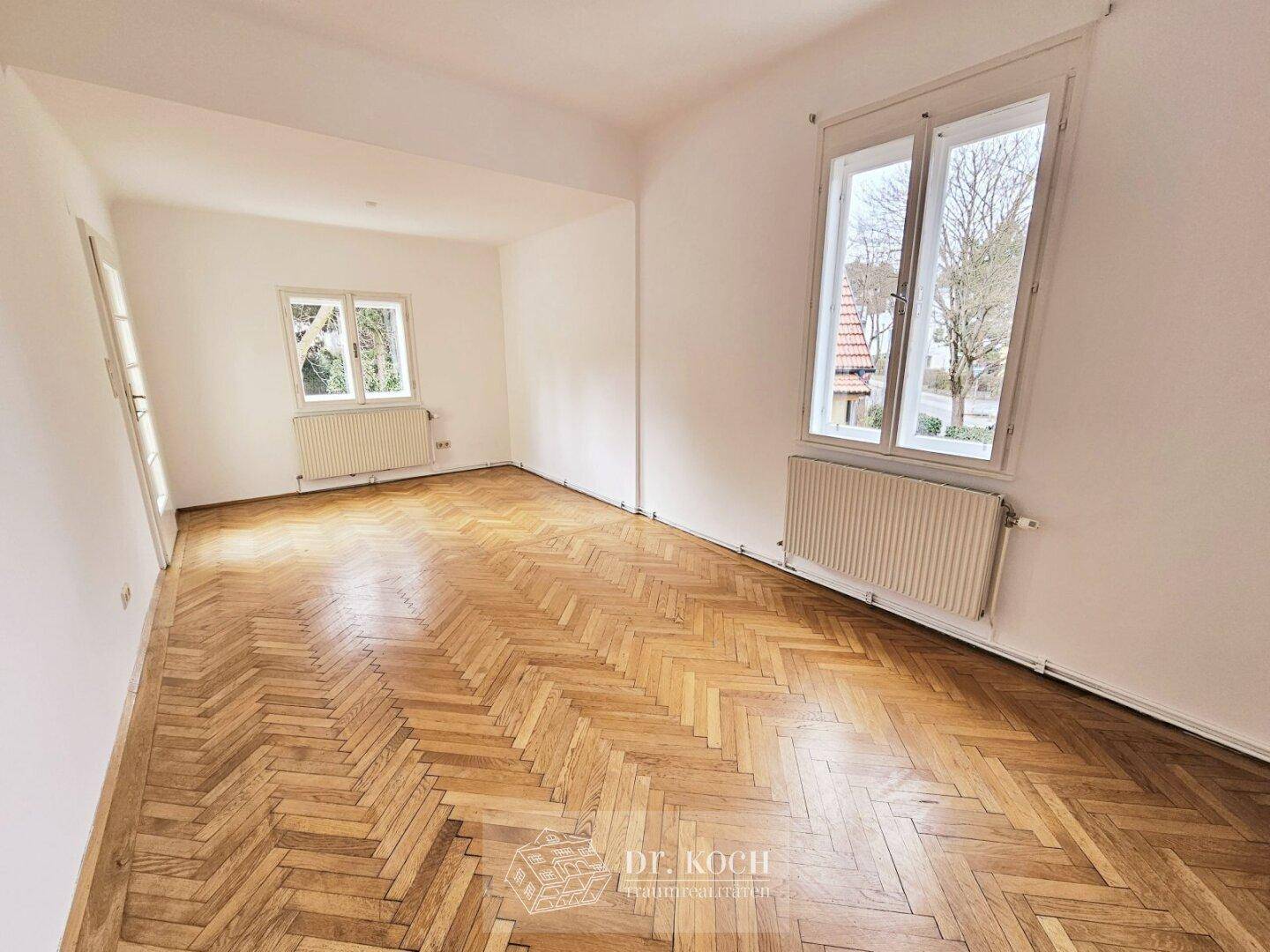 Heller Raum mit Fischgrätparkett und Blick auf die Straße durch große Fenster.