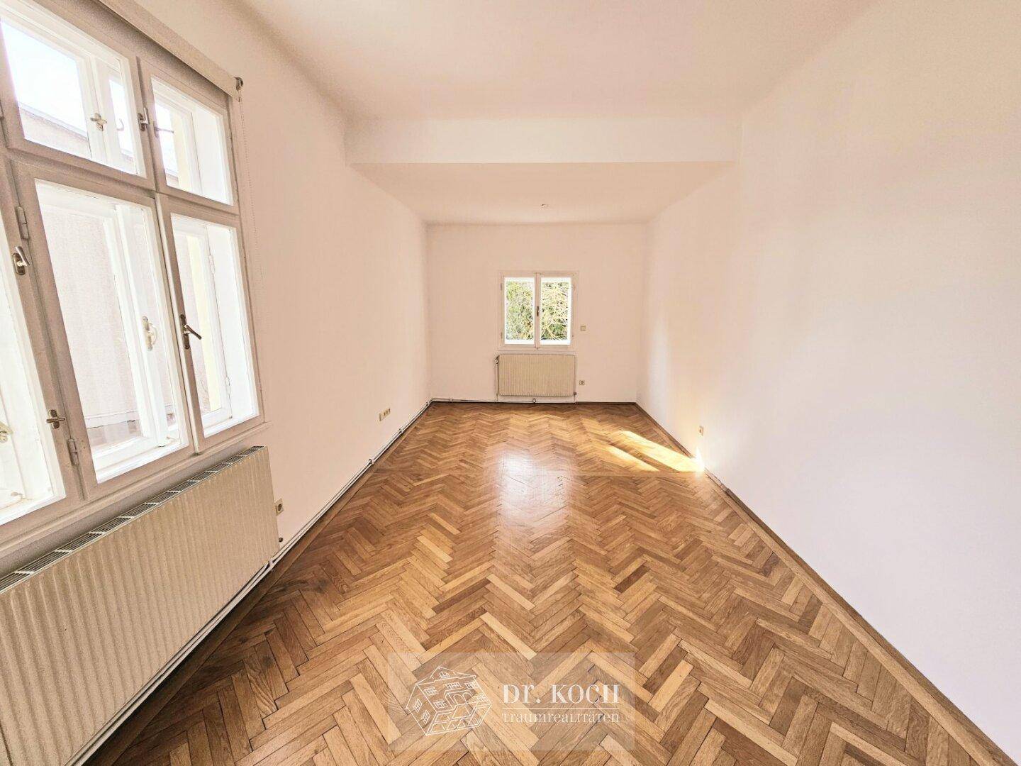 Langes, helles Zimmer mit Fischgrätparkett und Fenstern, die viel Licht spenden.