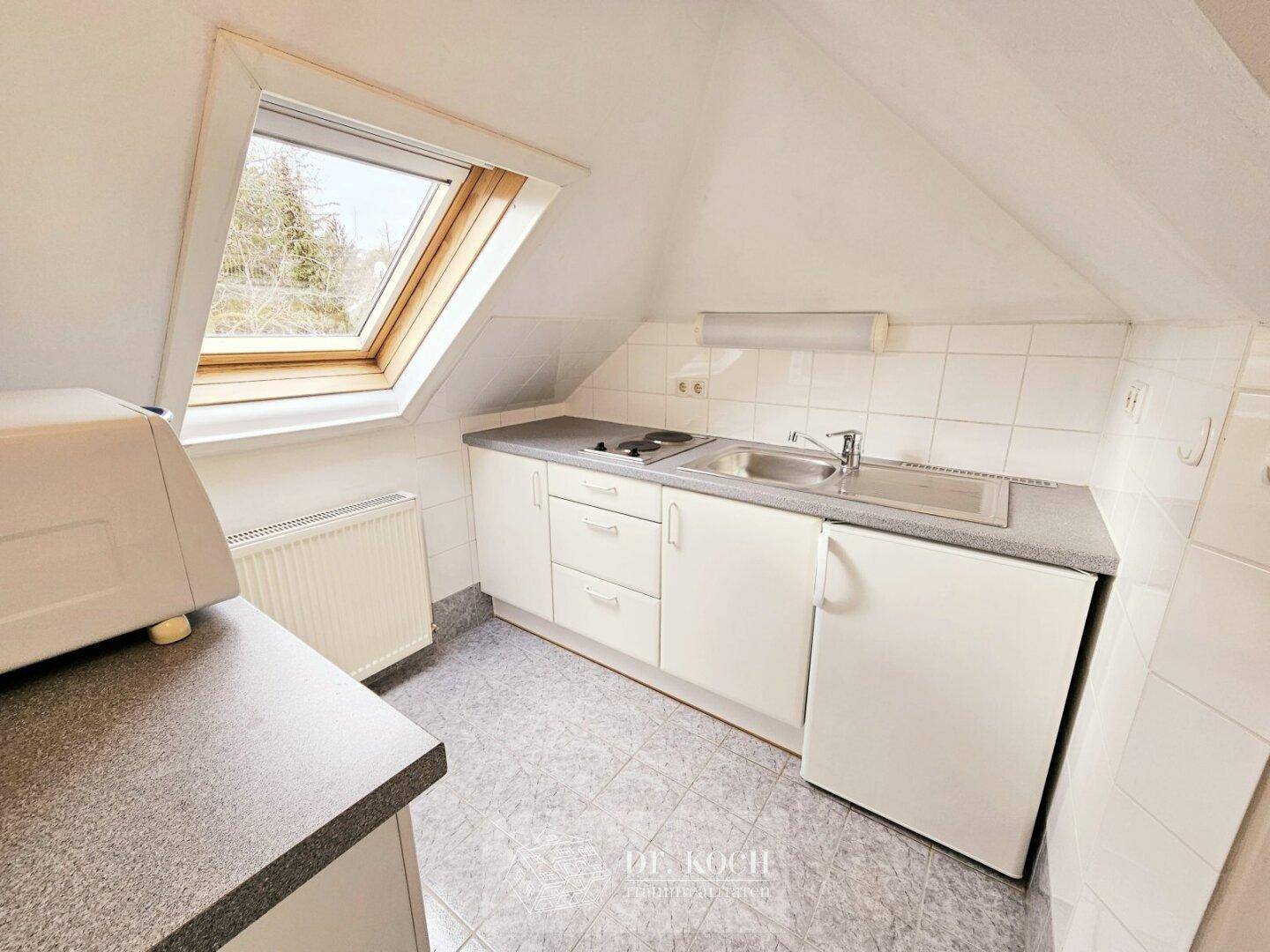 Kleine Küchenzeile im Dachgeschoss mit Spüle, Kochfeld und Kühlschrank unter einem Dachfenster.