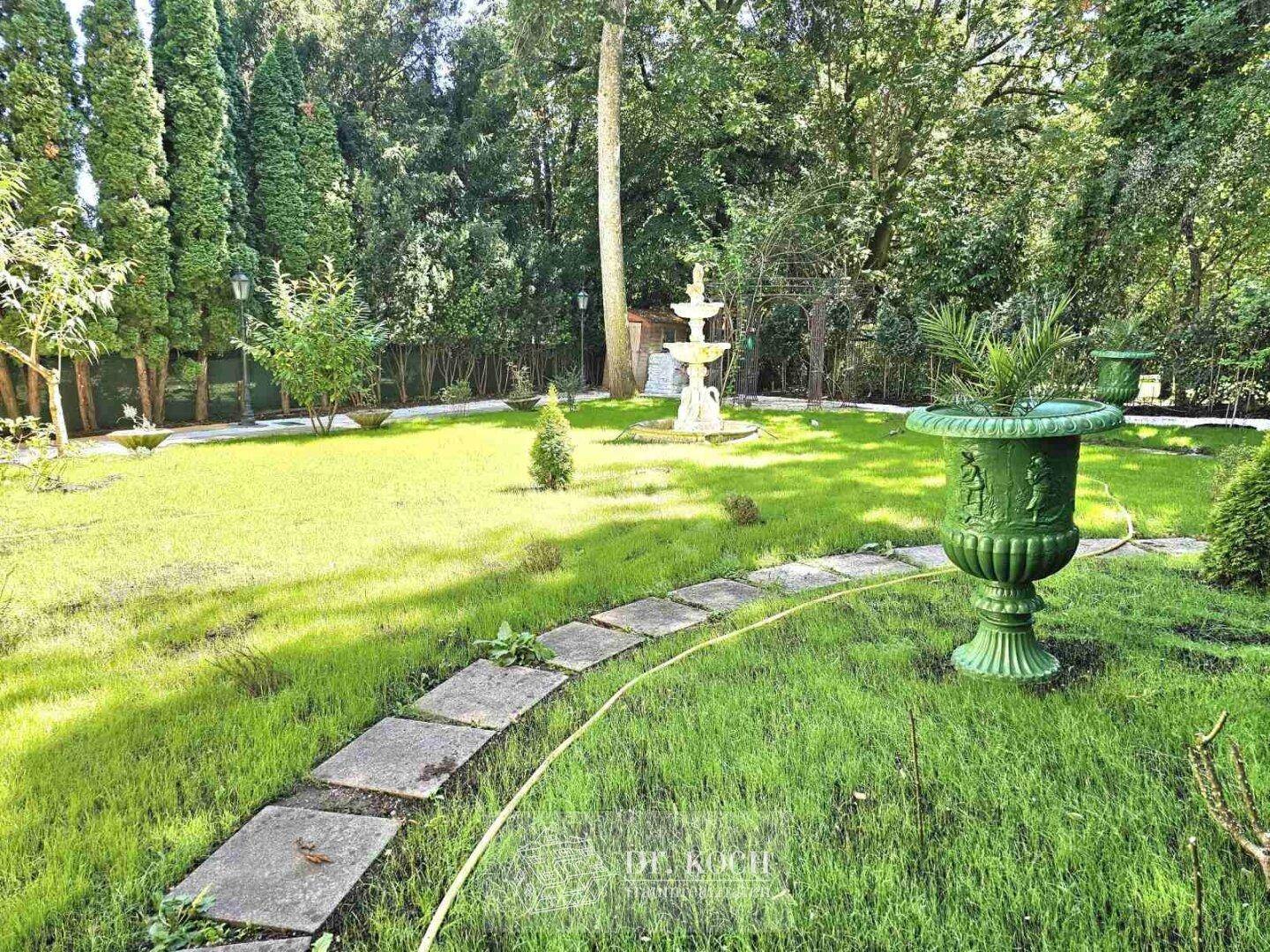 Weitläufiger, gepflegter Gartenbereich mit Rasenflächen, Bäumen und einem dekorativen Brunnen im Hintergrund.