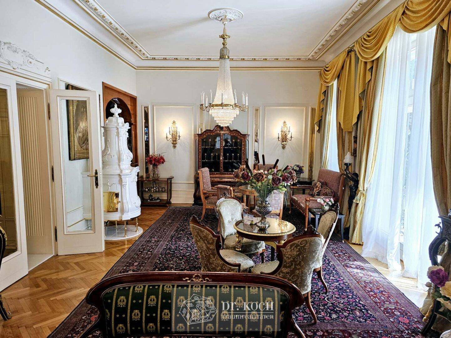 Großzügiges Wohnzimmer mit hohen Decken, Stuckverzierungen, Kachelofen und Zugang zu großen Fenstern.