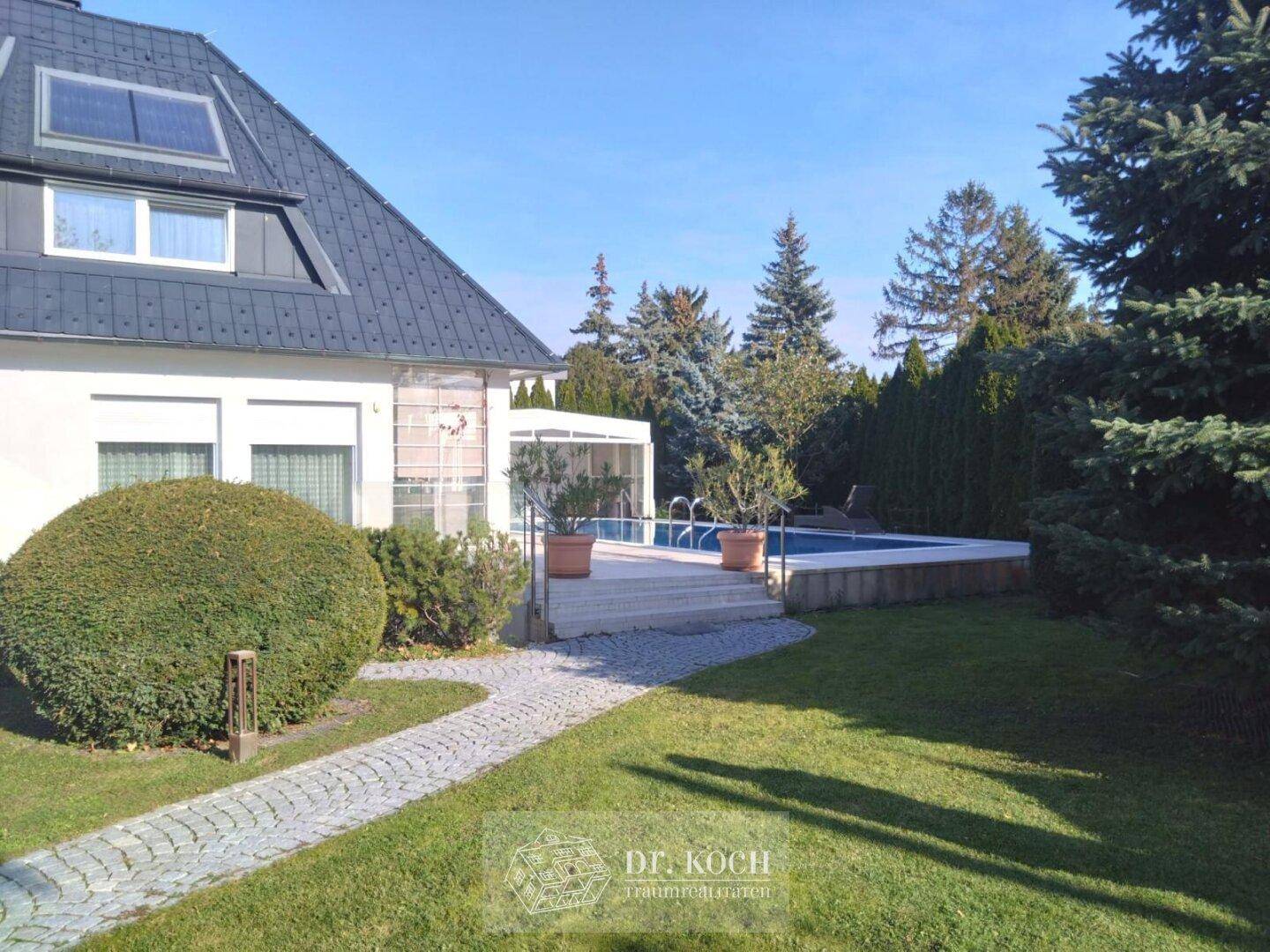 Hausfassade mit Gartenbereich und Blick auf den dahinterliegenden Swimmingpool.