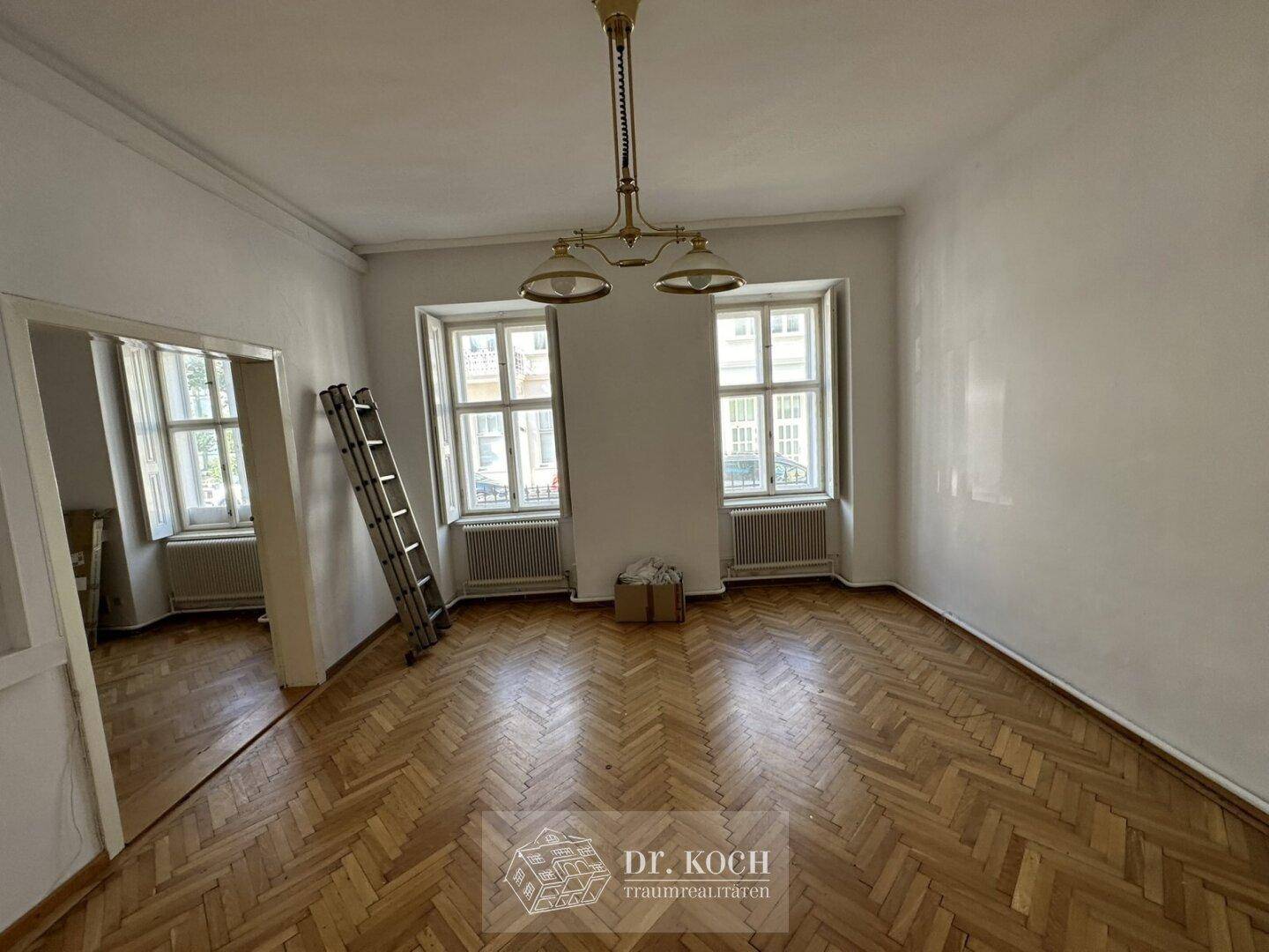 Geräumiges Wohnzimmer mit Fischgrätparkett, hohen Decken und zwei großen Fenstern.