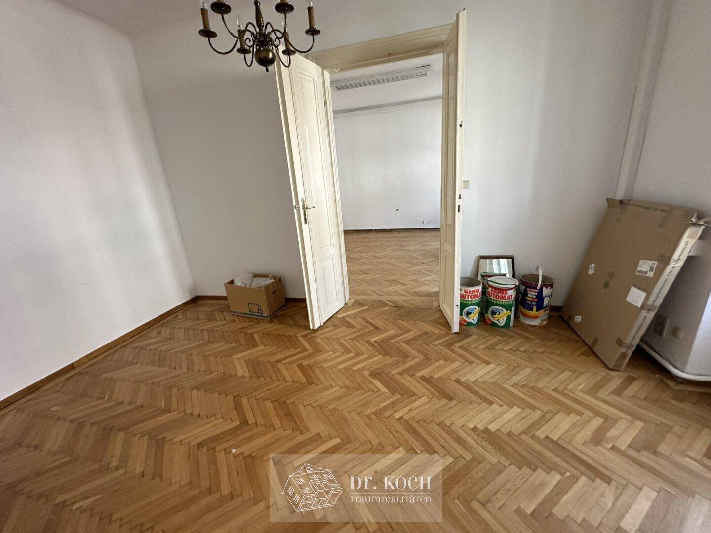 Geräumiges Zimmer mit Fischgrätparkett und einer Doppeltür zu einem weiteren Raum.