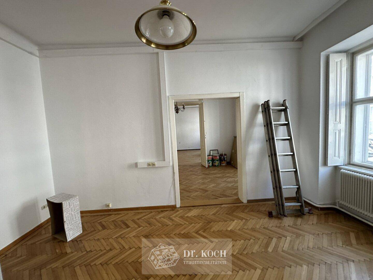 Helles Zimmer mit Fischgrätparkett, hohen Decken und einem großen Fenster.