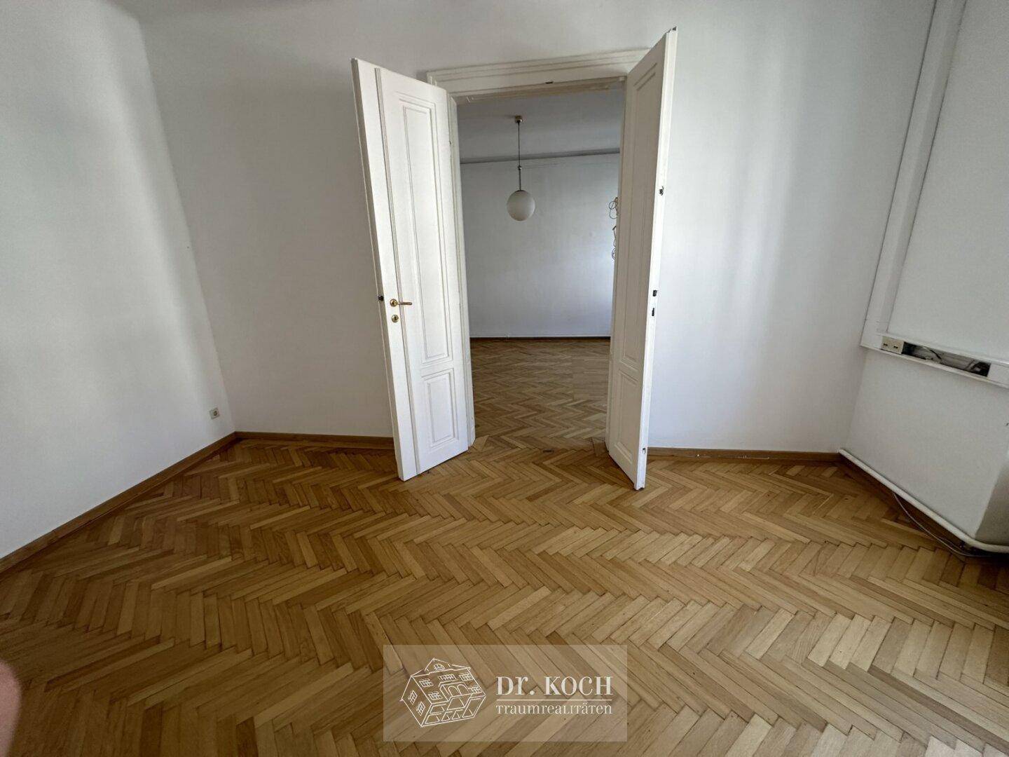 Helles Zimmer mit Fischgrätparkett und einer weit geöffneten Tür zu einem weiteren Raum.