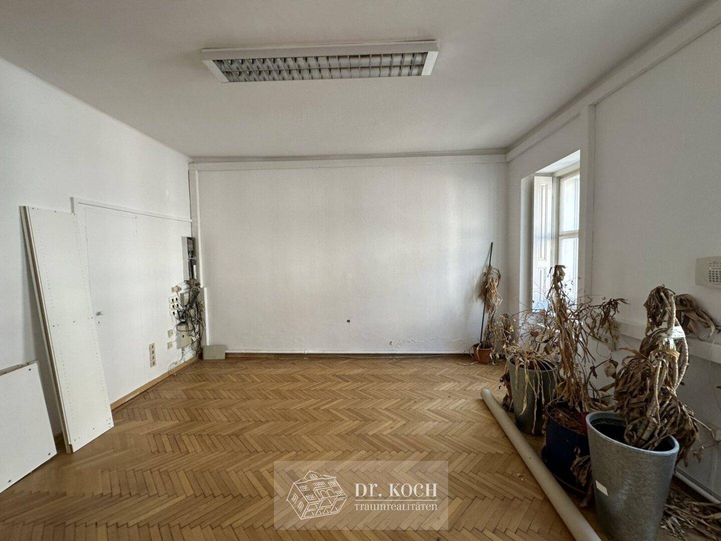 Großer Raum mit Fischgrätparkett und einer Deckenleuchte, renovierungsbedürftig.