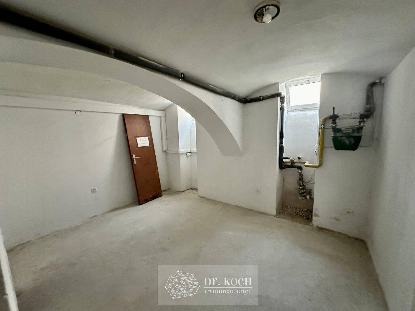 Kellerraum mit Fenster, Gasleitungen und einem Durchgang zu einem weiteren Bereich.