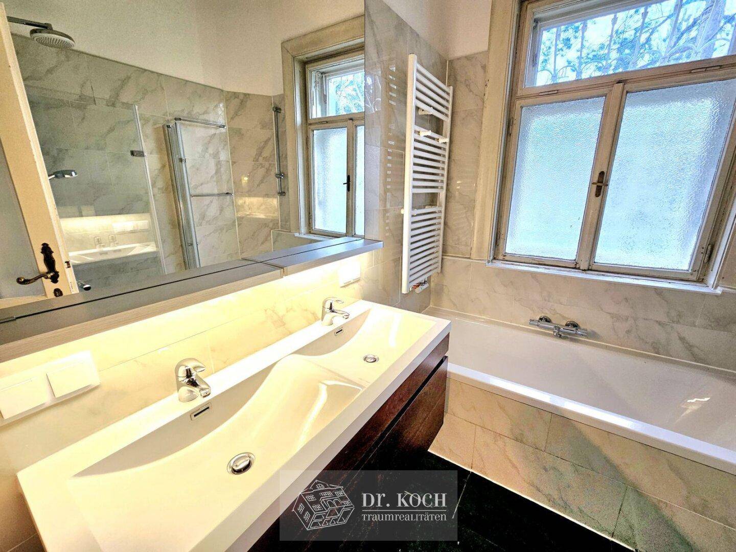 Elegantes Badezimmer mit Doppelwaschbecken, Badewanne und zwei Fenstern für viel Tageslicht.