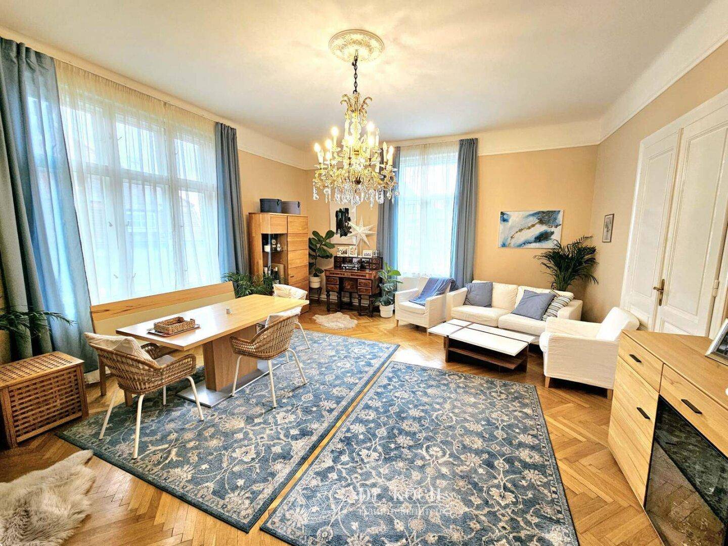 Eleganter Wohn-Essbereich mit Kronleuchter, Parkettboden und großen Fenstern mit blauen Vorhängen.