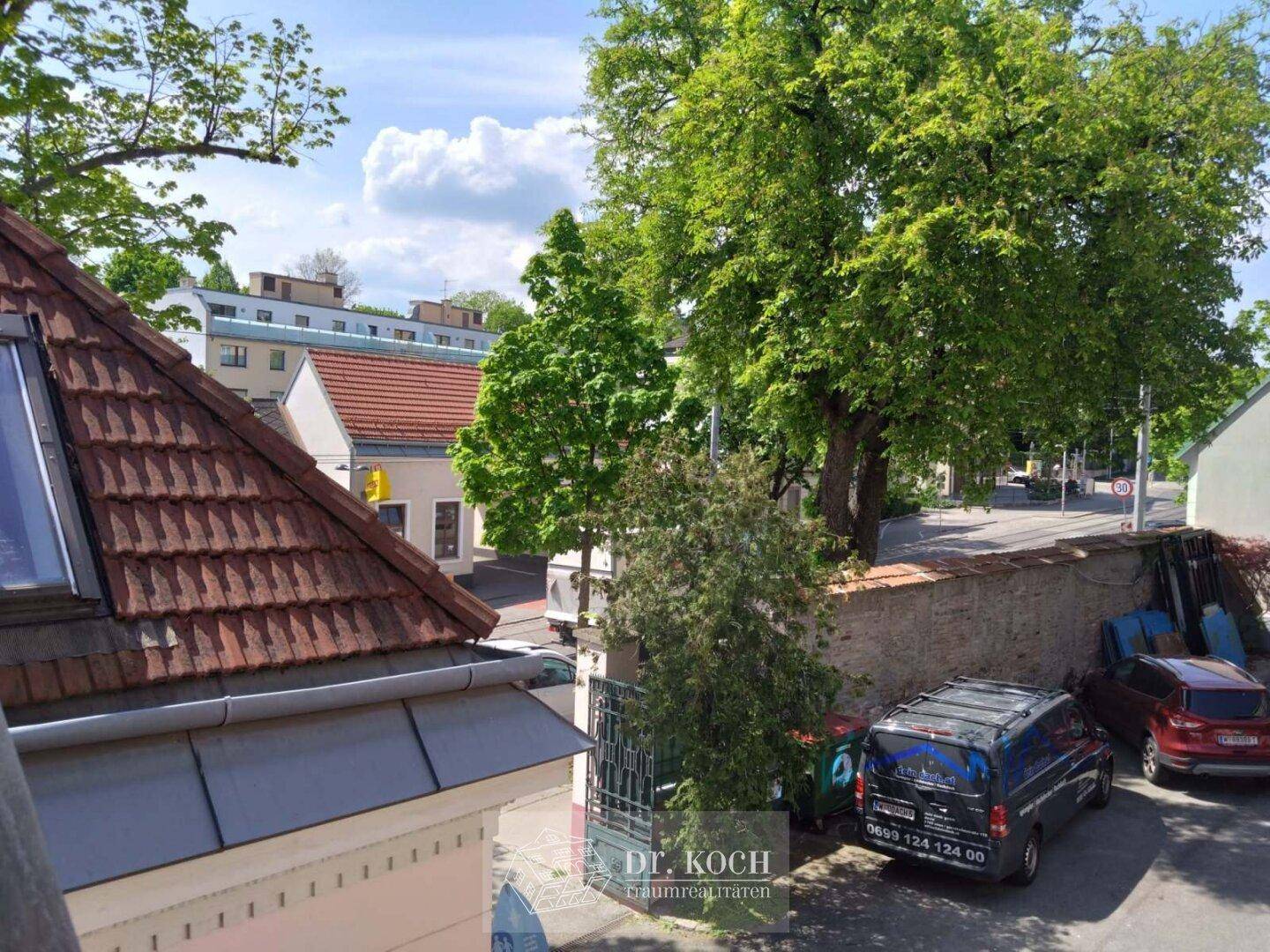 Blick von oben auf eine belebte Straße mit parkenden Fahrzeugen und üppigen Bäumen, umgeben von Gebäuden.