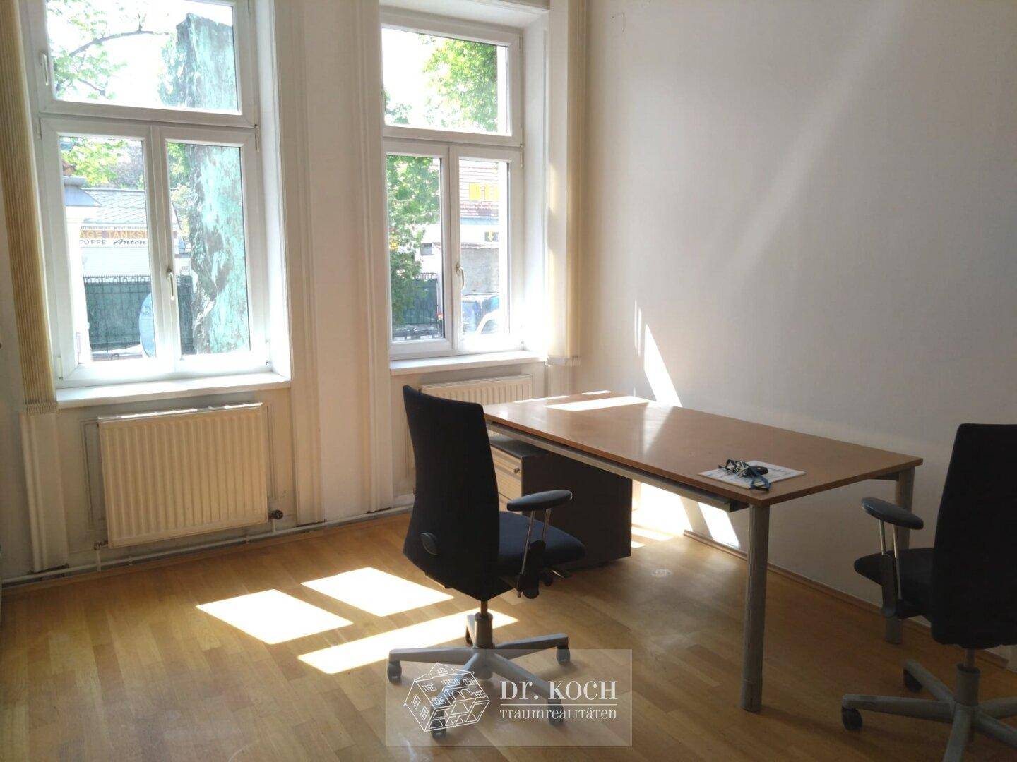 Helles Büro mit zwei großen Fenstern, Holzboden und einem Schreibtisch mit Bürostühlen, bietet angenehme Arbeitsatmosphäre.