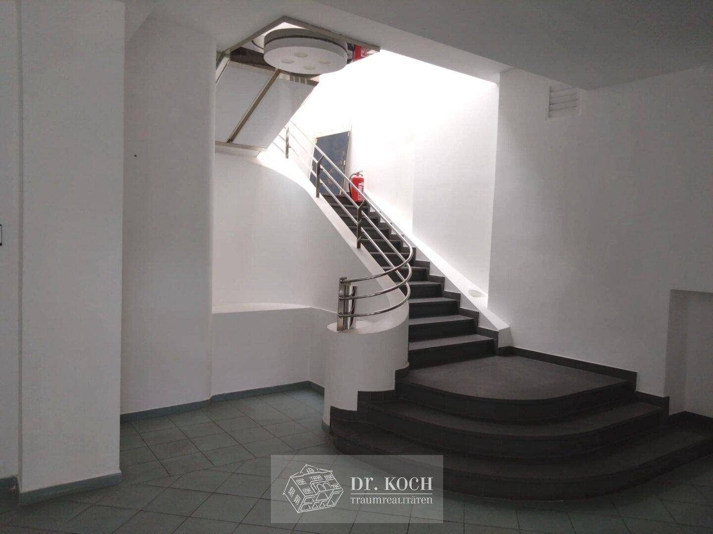 Modernes Treppenhaus mit dunklen Stufen, elegantem Metallgeländer und viel Tageslicht von oben.
