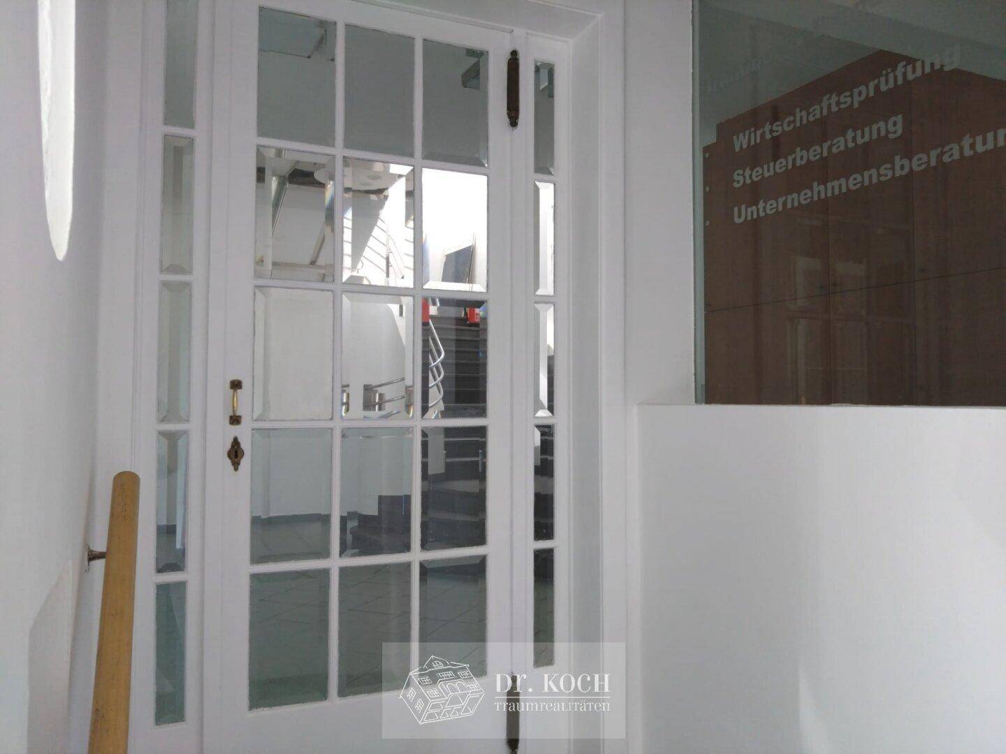 Weiße Glastür mit Sprossen in einem hellen Flur, der zu einem Treppenhaus und weiteren Räumen führt.