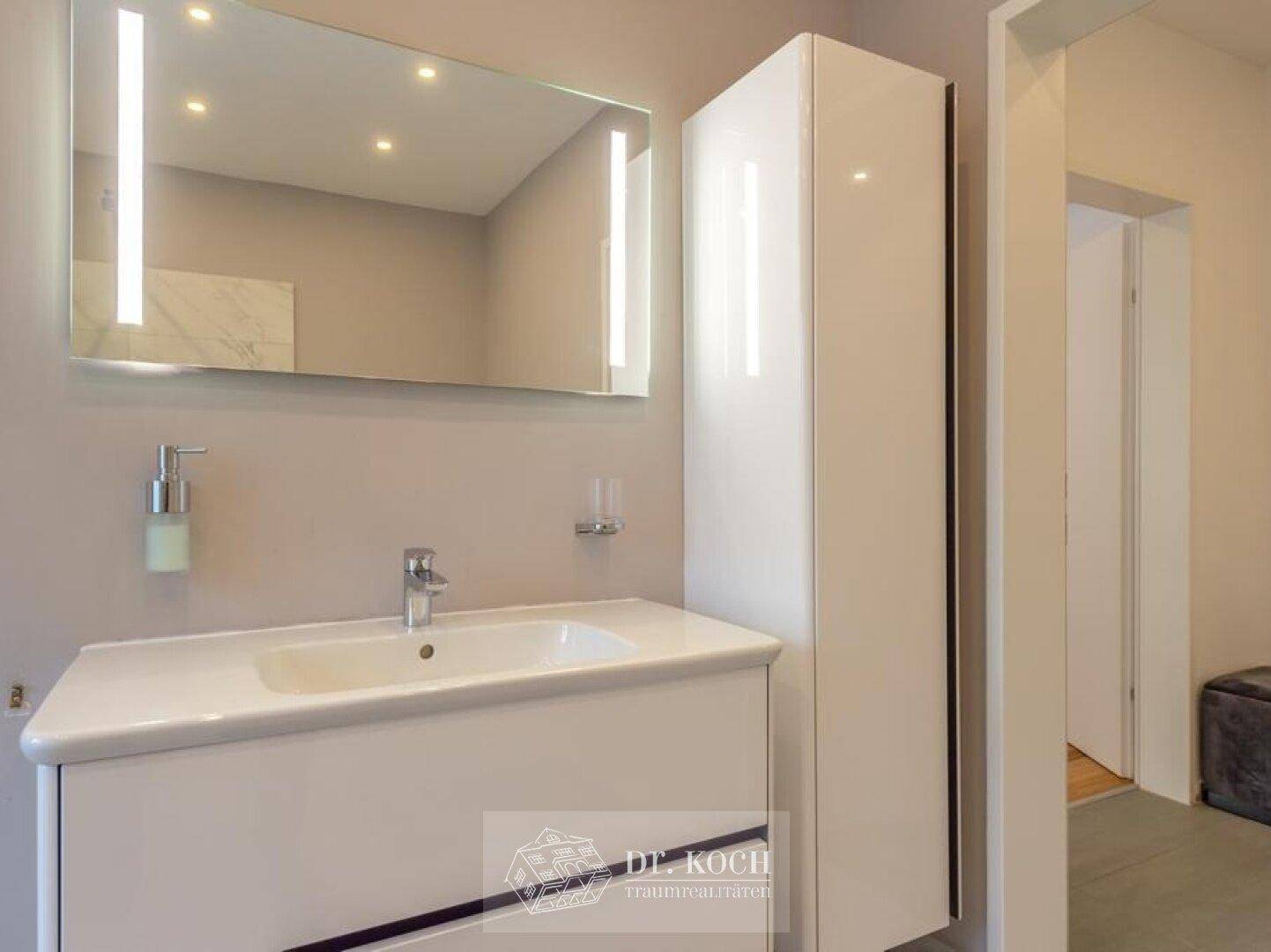 Modernes Badezimmer mit beleuchtetem Spiegel, Einzelwaschbecken und weißem Schrank.