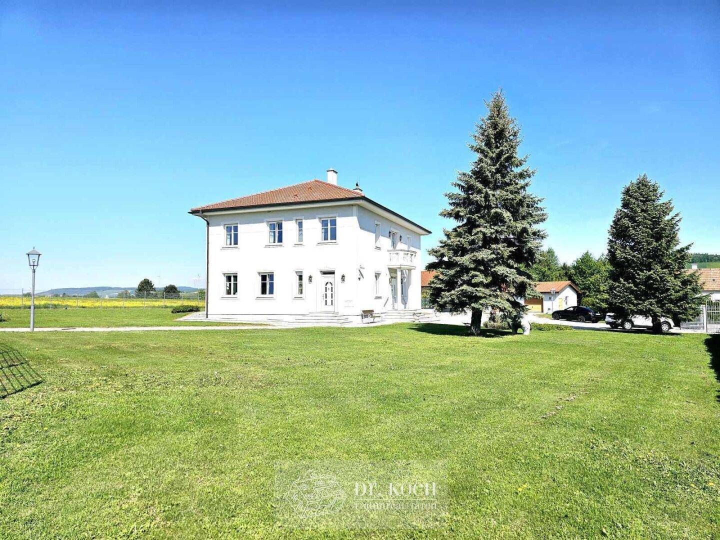 Elegantes Landhaus mit weitläufiger Rasenfläche und hohen Nadelbäumen unter strahlend blauem Himmel.