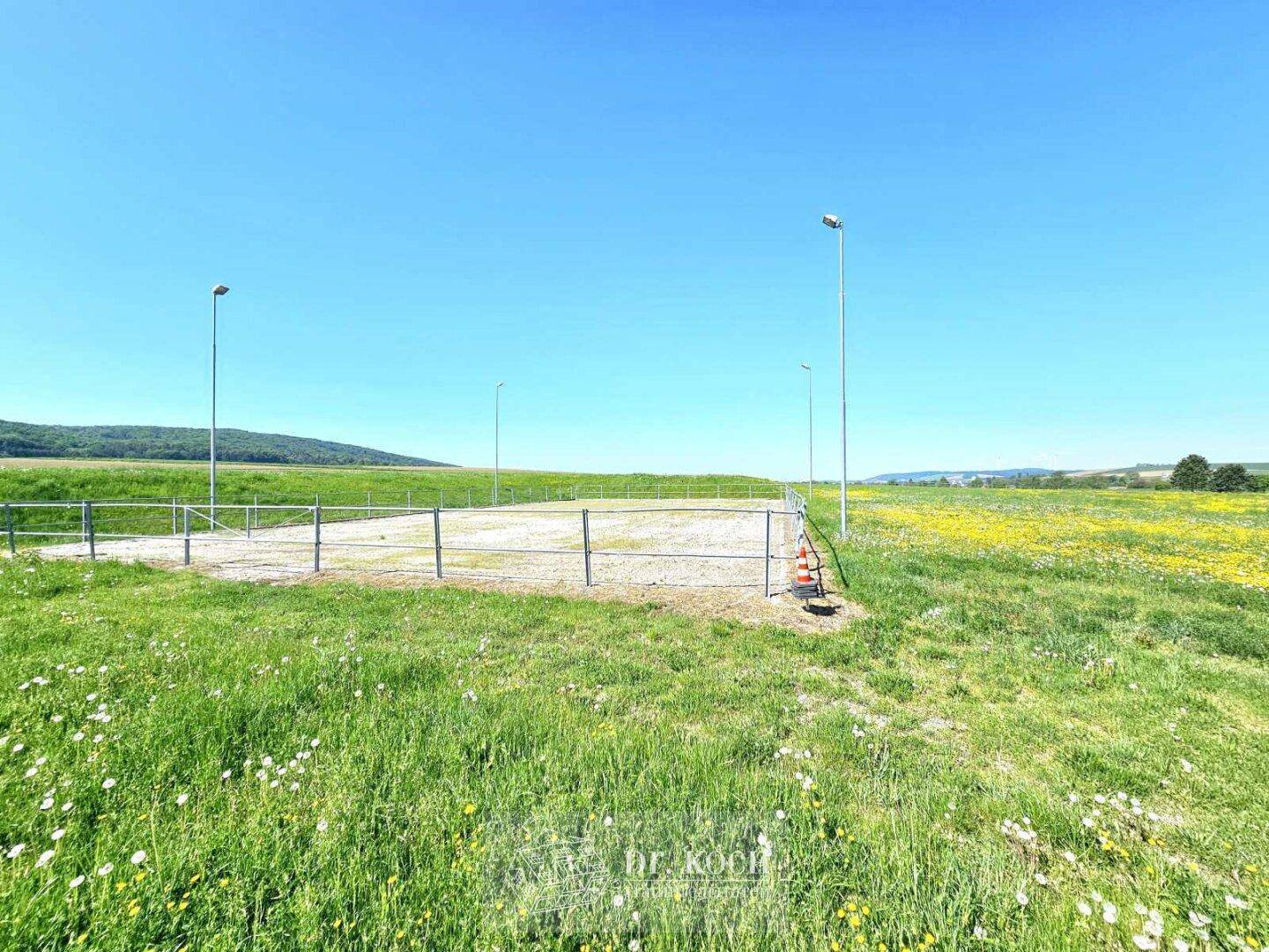 Outdoor-Sportplatz oder Reitplatz umgeben von grünen Wiesen und Hügeln unter blauem Himmel.