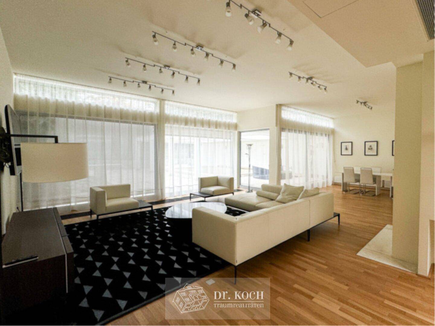 Heller, offener Wohnbereich mit großen Fenstern, modernem Sofa und Essbereich im Hintergrund.