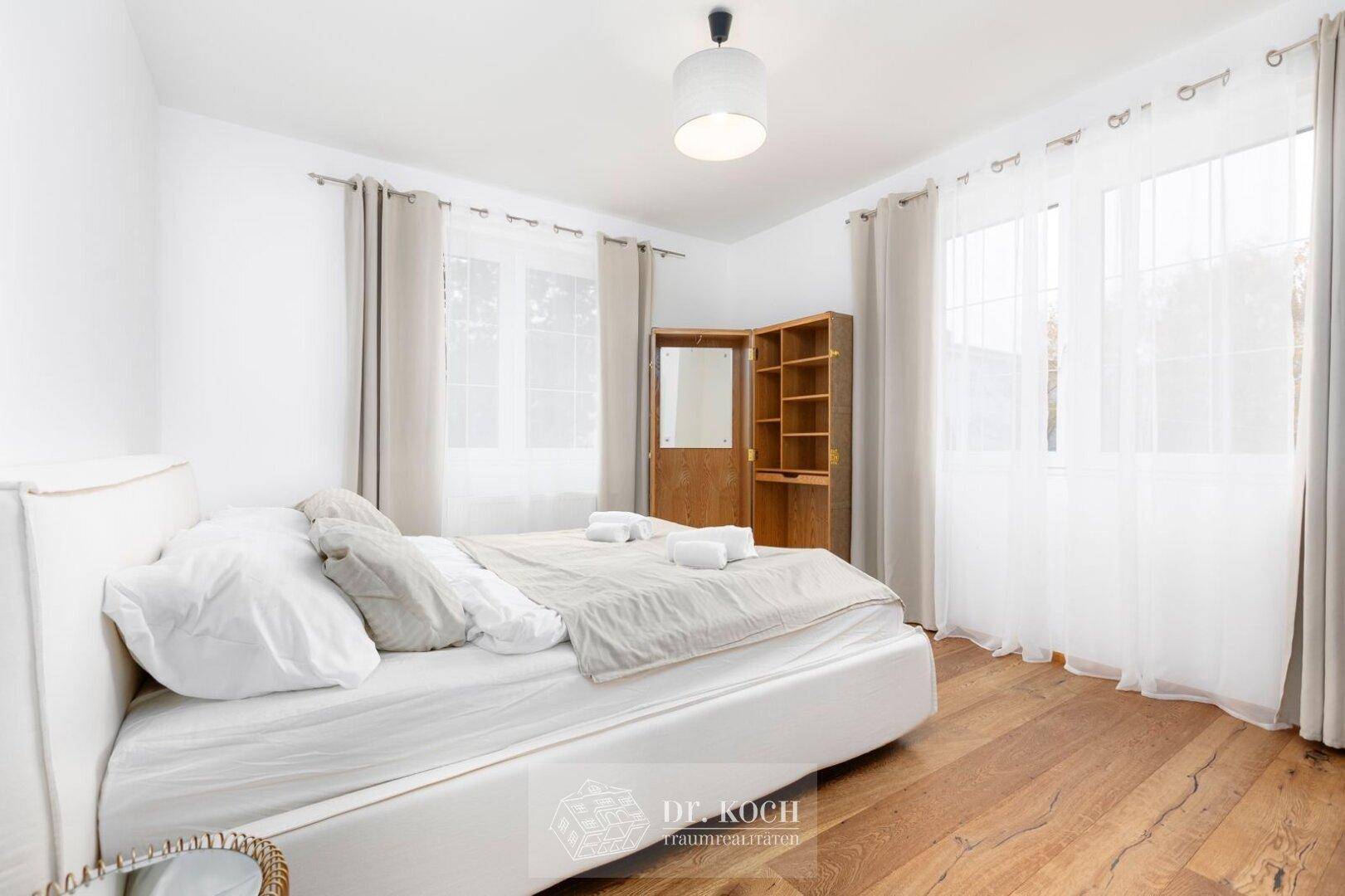 Helles Schlafzimmer mit Holzboden, weißem Bett und großen Fenstern, die viel Licht hereinlassen.