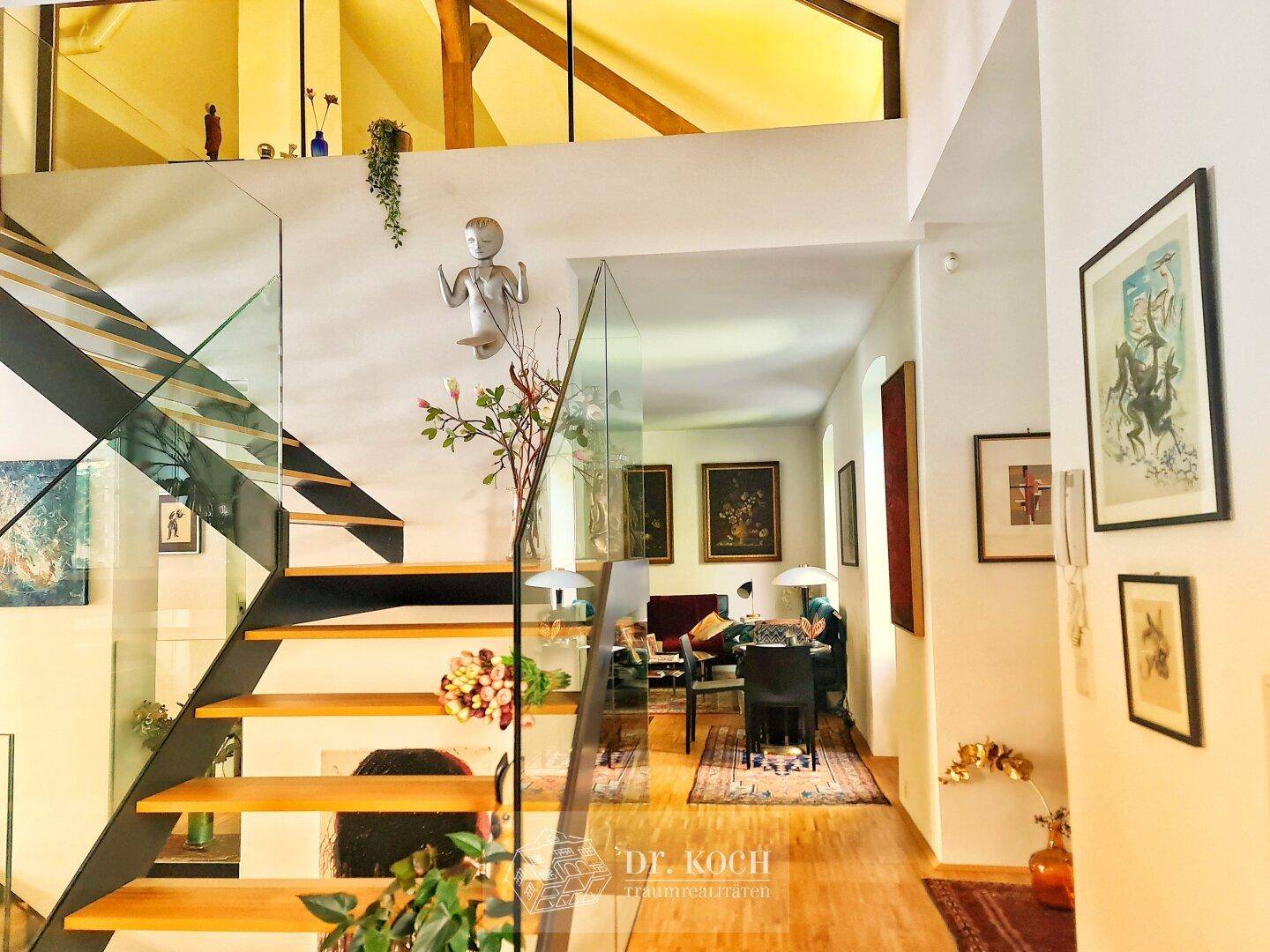 Beeindruckende moderne Treppe mit Glasgeländer und Blick auf den offenen Wohnbereich mit Holzbalken.