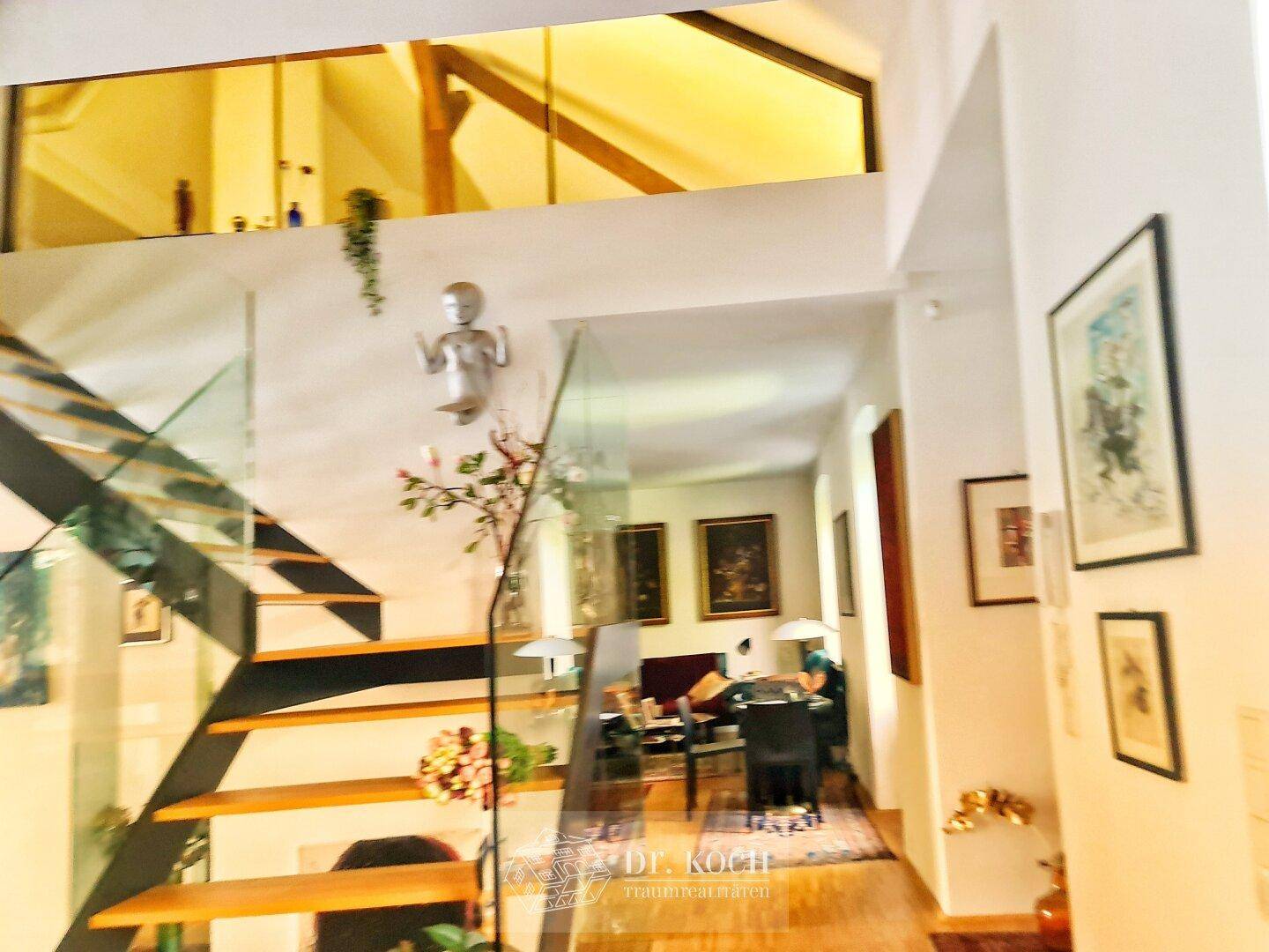 Beeindruckende moderne Treppe mit Glasgeländer und Blick auf den offenen Wohnbereich mit Holzbalken.