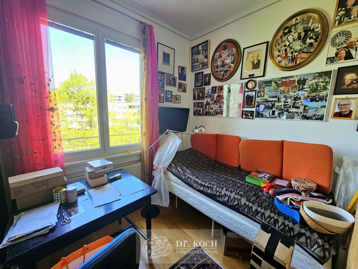Helles Zimmer mit Bett, Schreibtisch und vielen gerahmten Bildern an der Wand, mit Blick ins Grüne.