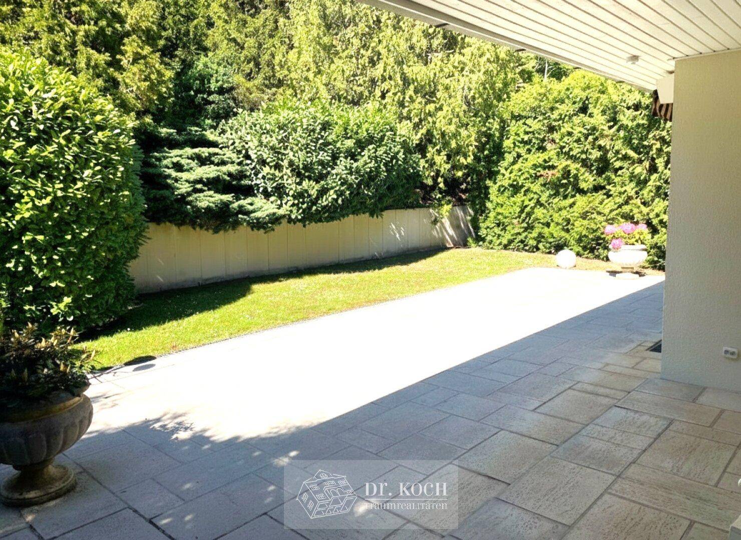 Sonnige Terrasse mit Steinplatten und angrenzender Rasenfläche, umgeben von Hecken.