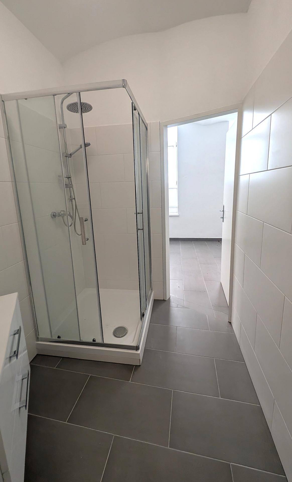 Helles Badezimmer mit einer modernen Duschkabine und grauen Bodenfliesen, die zu einem weiteren Raum führen.