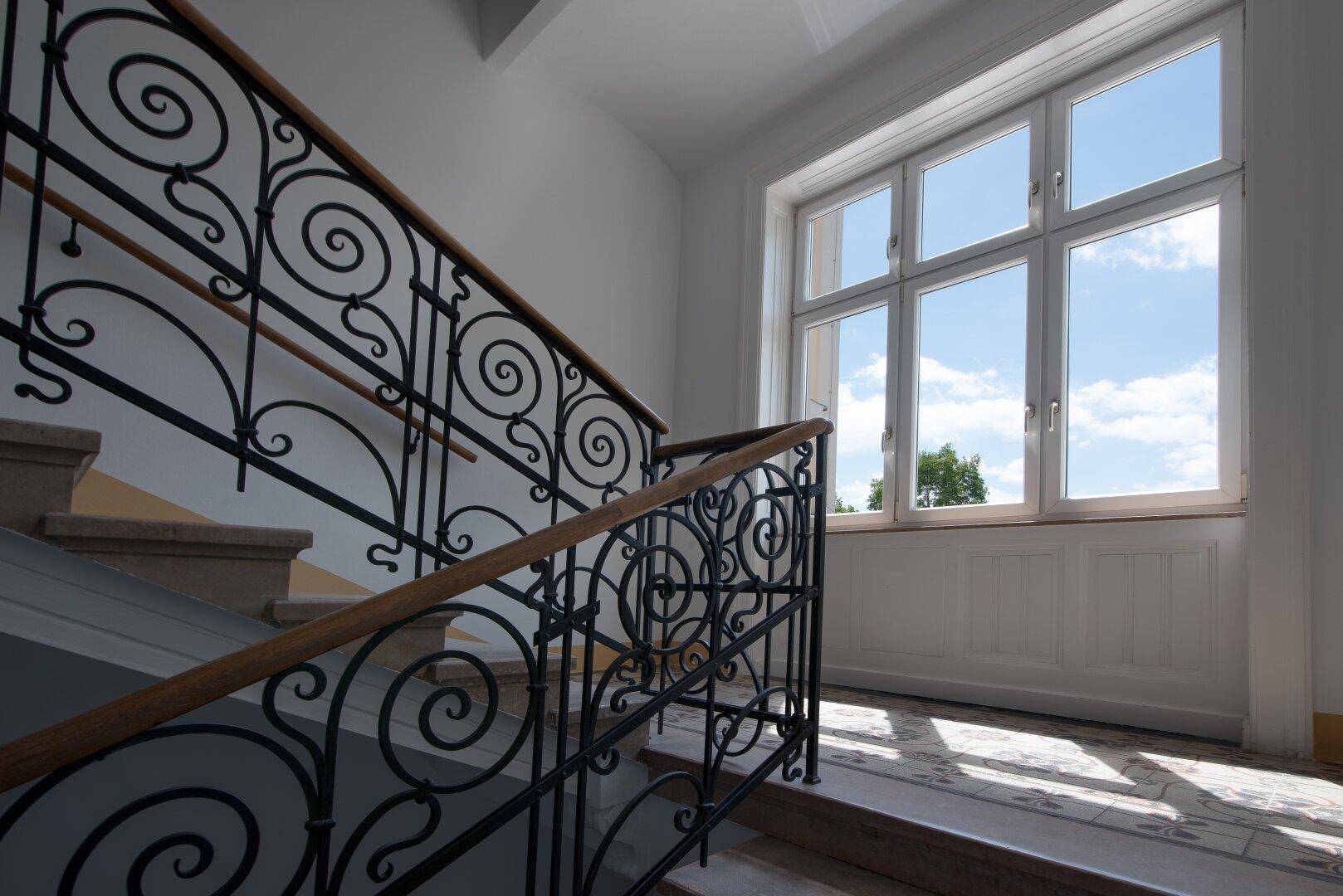 Elegantes Treppenhaus mit schmiedeeisernem Geländer, Holzhandlauf und großem Fenster für viel Tageslicht.