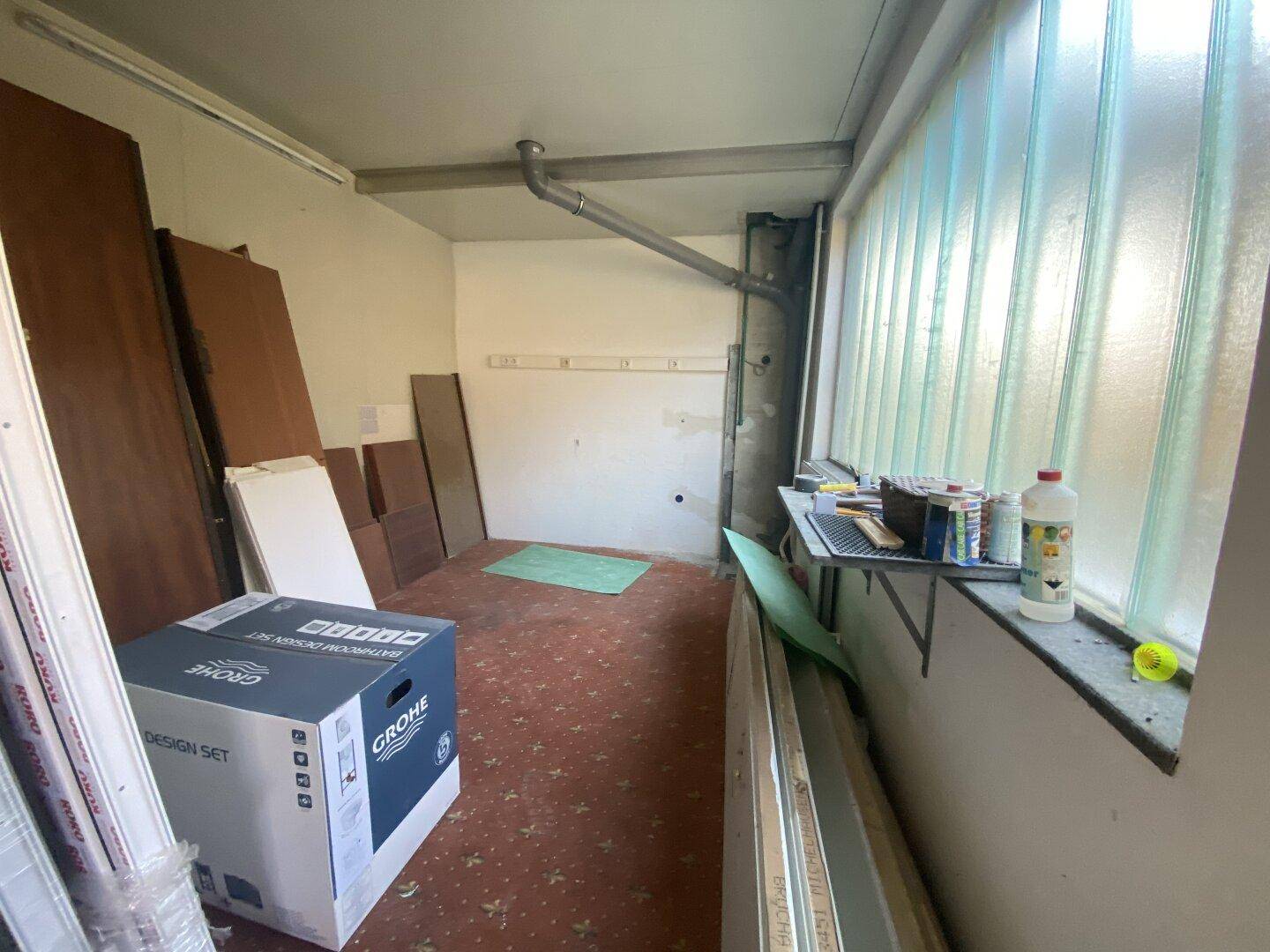 Kleinerer Nebenraum mit Glasbausteinfenster, sichtbaren Rohren und Teppichboden, ideal als Abstellraum.