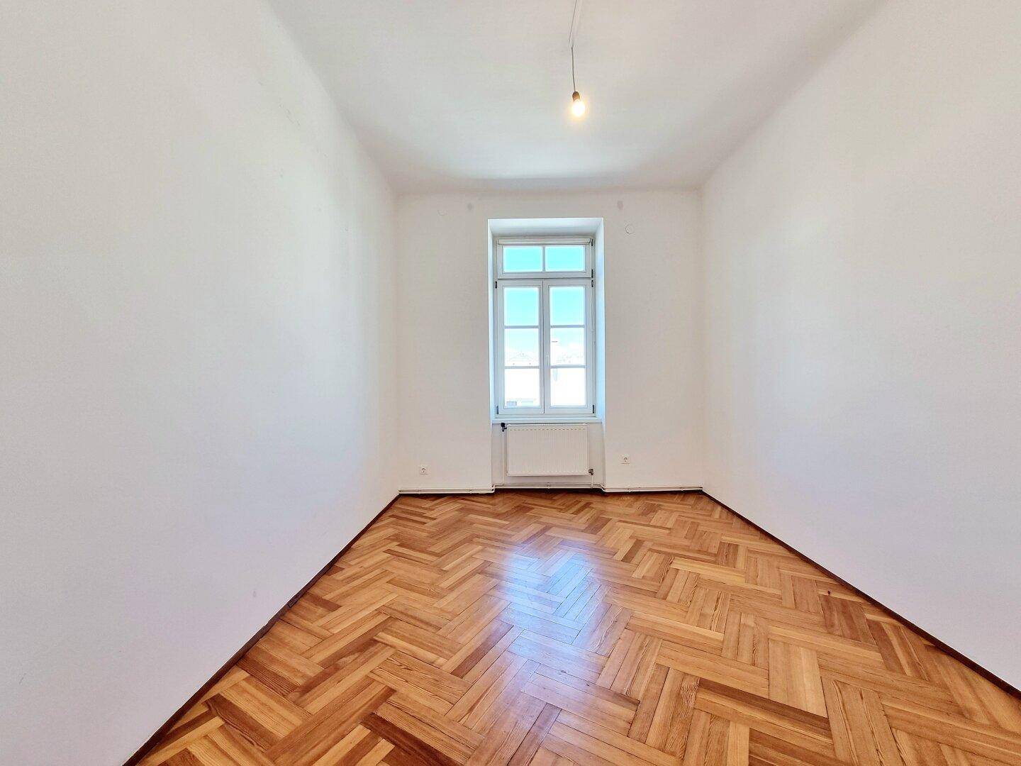 Helles Zimmer mit Fischgrätparkett und einem großen Fenster, geeignet als Schlafzimmer.