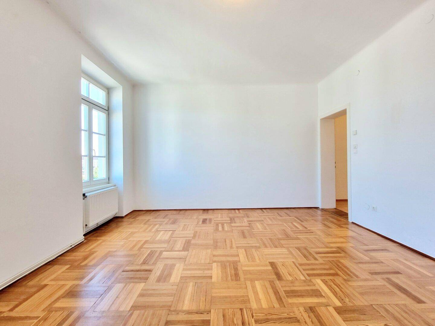 Geräumiges, helles Zimmer mit klassischem Fischgrätparkett und einem großen Fenster.