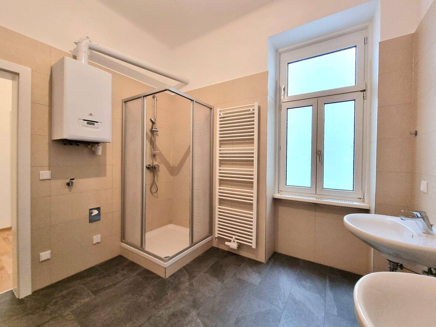 Helles Badezimmer mit Duschkabine, Waschbecken, Handtuchheizkörper und Fenster.