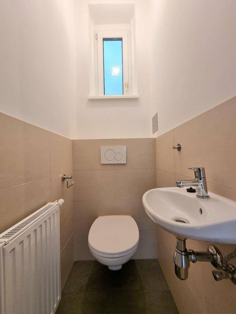 Separates WC mit wandhängender Toilette, kleinem Waschbecken und Fenster.