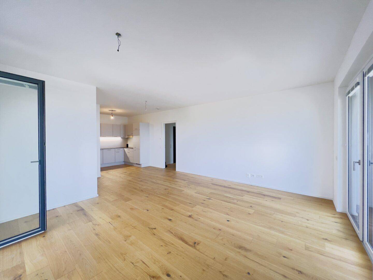 Weitläufiger Wohnbereich mit Holzboden, weißen Wänden und Blick auf die offene Küche.