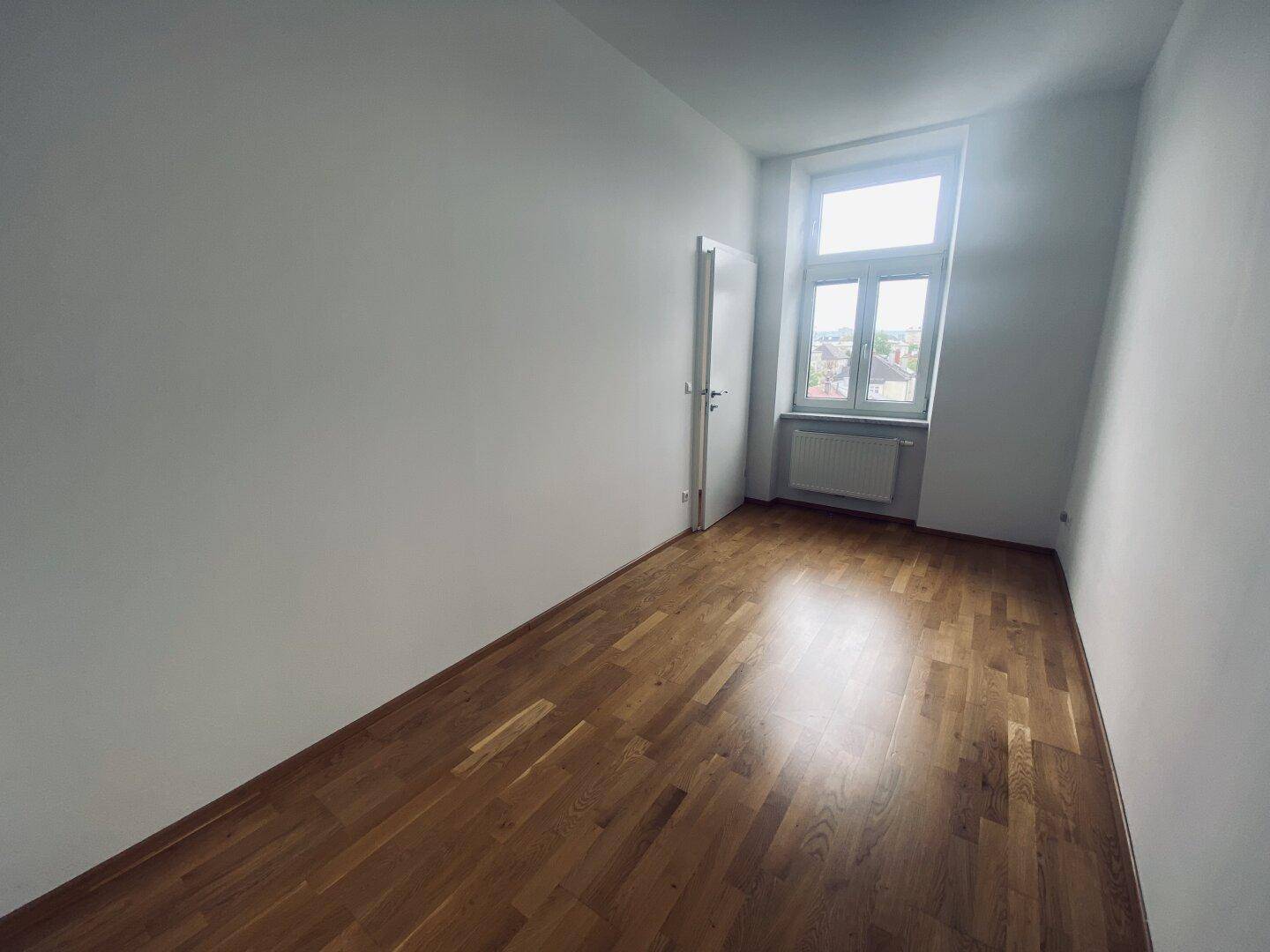 Leeres Zimmer mit warmem Parkettboden, weißen Wänden und einem Fenster, das viel Licht hereinlässt.