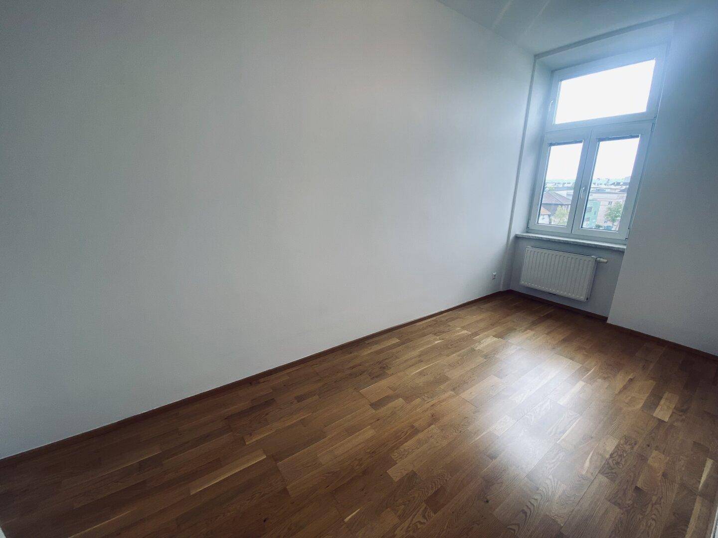 Helles und leeres Zimmer mit Parkettboden, weißen Wänden und einem Fenster mit Blick nach draußen.