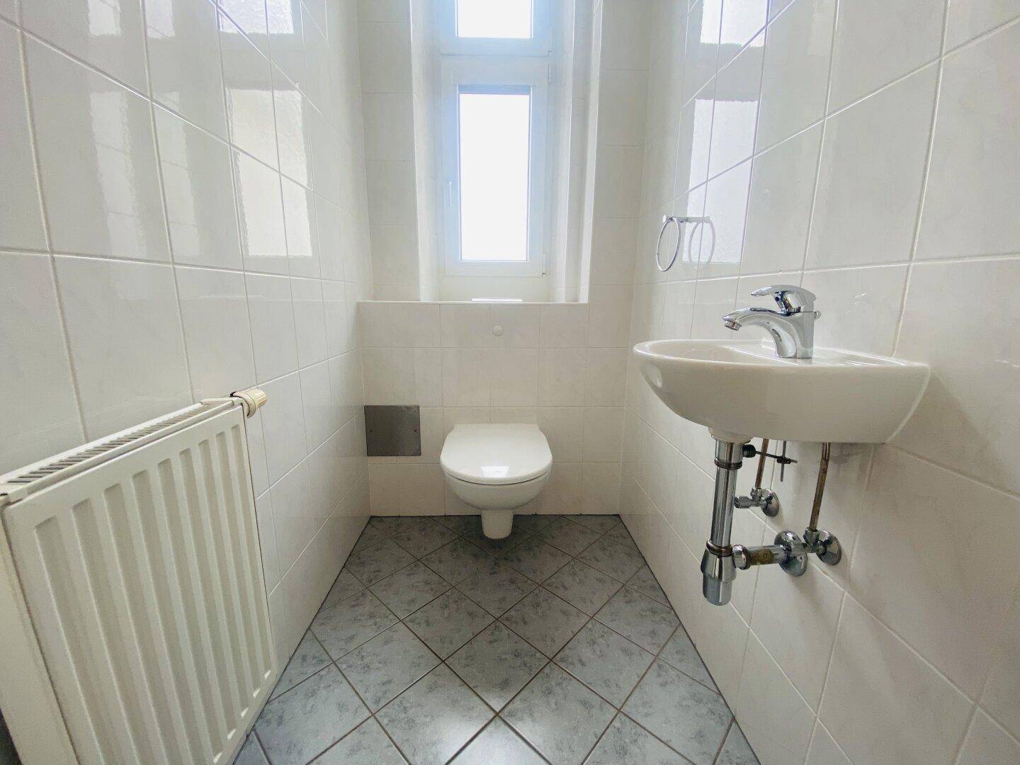 Kleines Gäste-WC mit weißen Fliesen, Toilette, Waschbecken, Fenster und Heizkörper.