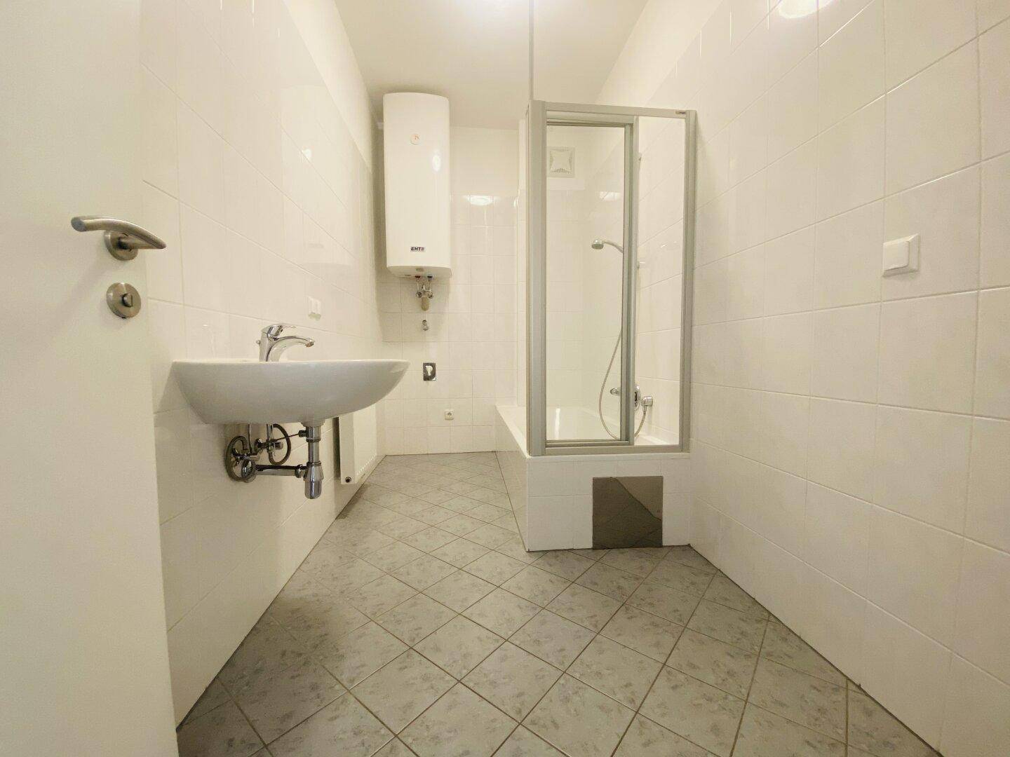 Helles Badezimmer mit weißen Fliesen, Waschbecken, Duschkabine und einem Warmwasserboiler.