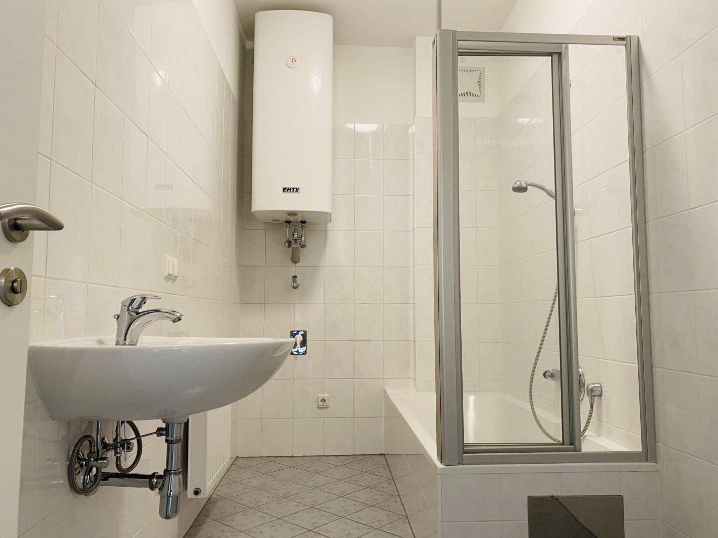 Funktionales Badezimmer mit weißen Fliesen, Waschbecken, Duschkabine und einem Warmwasserboiler.