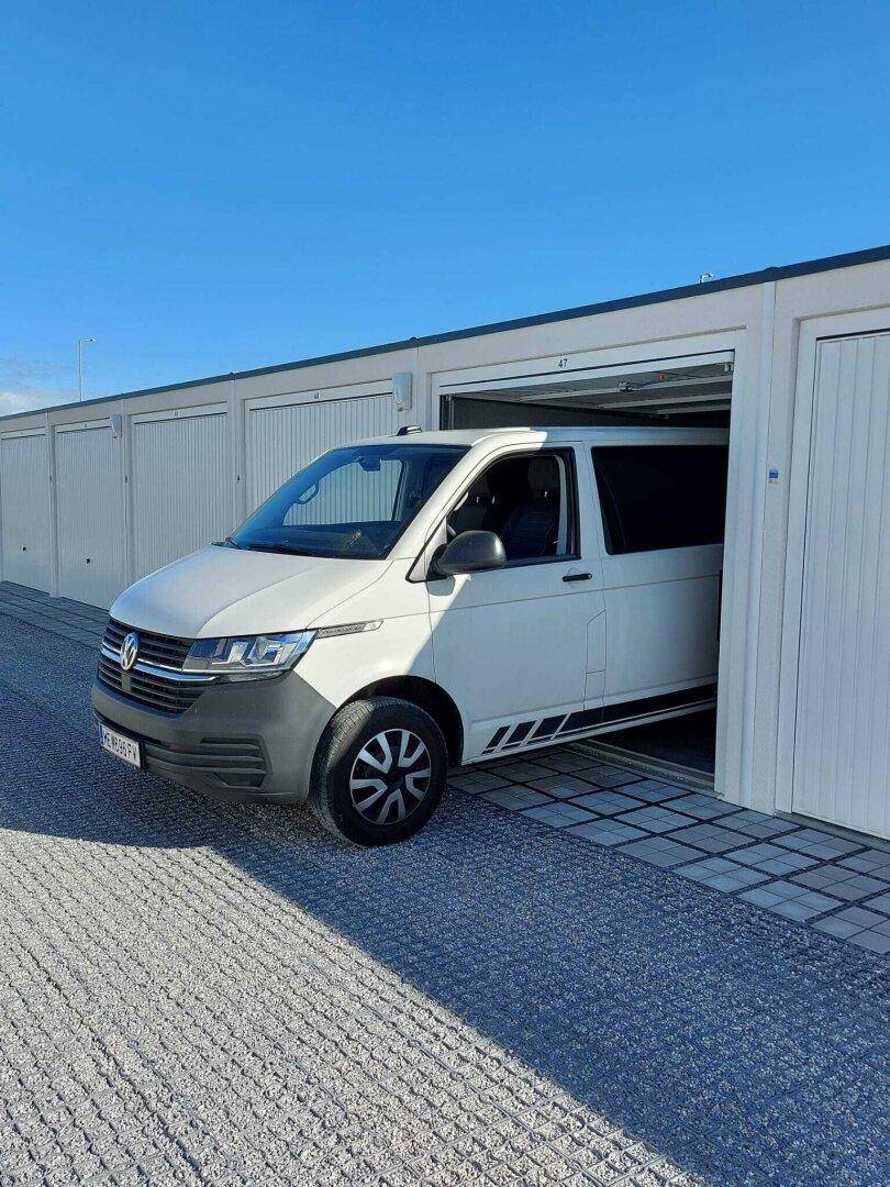 Ein weißer Volkswagen Transporter fährt aus einer modernen Garagenbox unter strahlend blauem Himmel heraus.