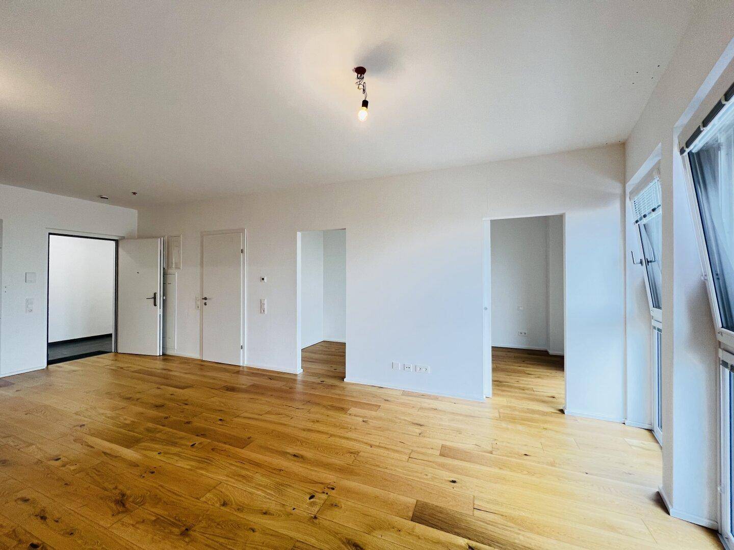 Geräumiges, helles Wohnzimmer mit glänzendem Parkettboden und mehreren Türen zu angrenzenden Räumen.