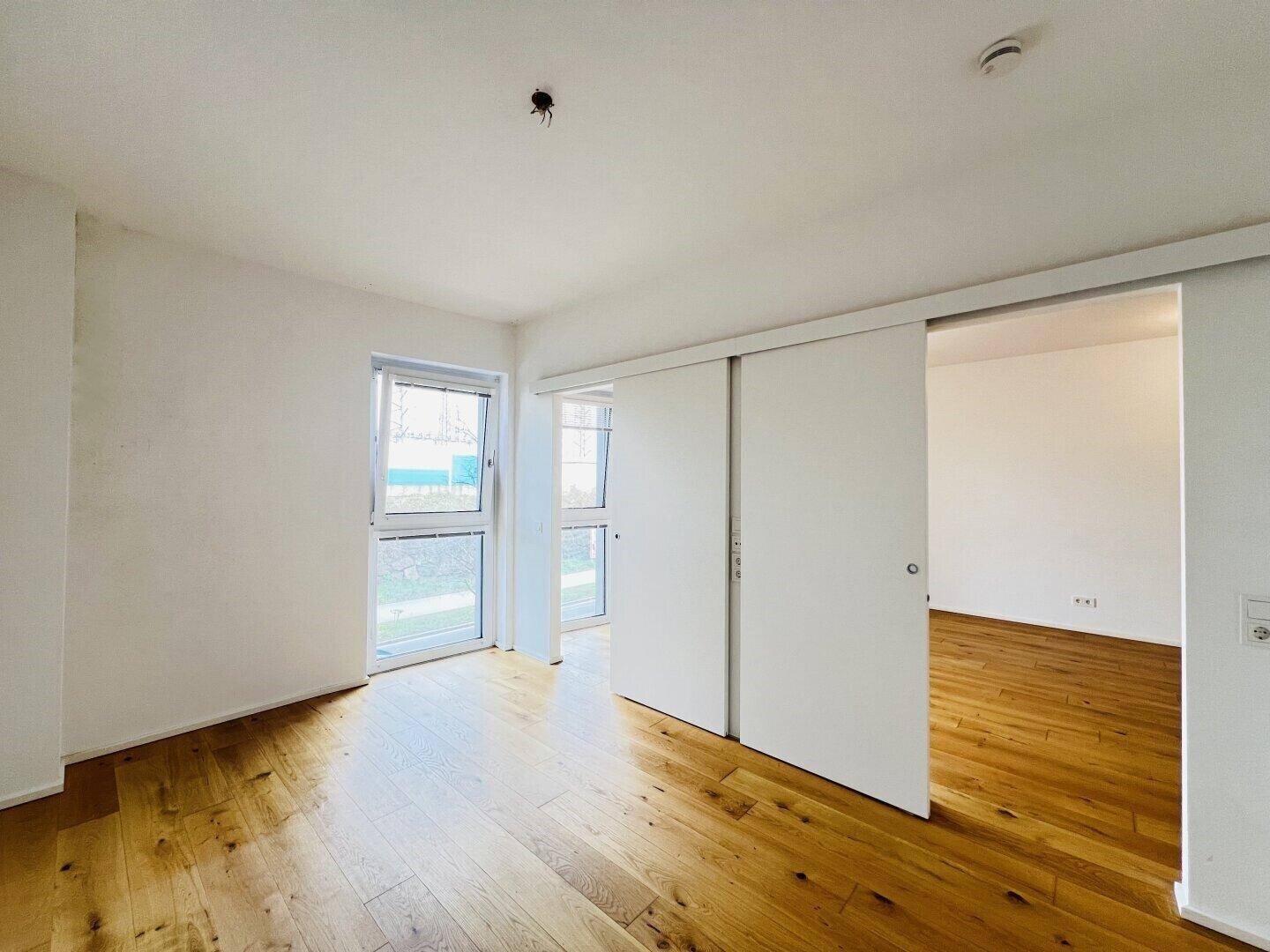 Helles Zimmer mit Holzboden, zwei Fenstern und einer modernen Schiebetür zu einem weiteren Raum.