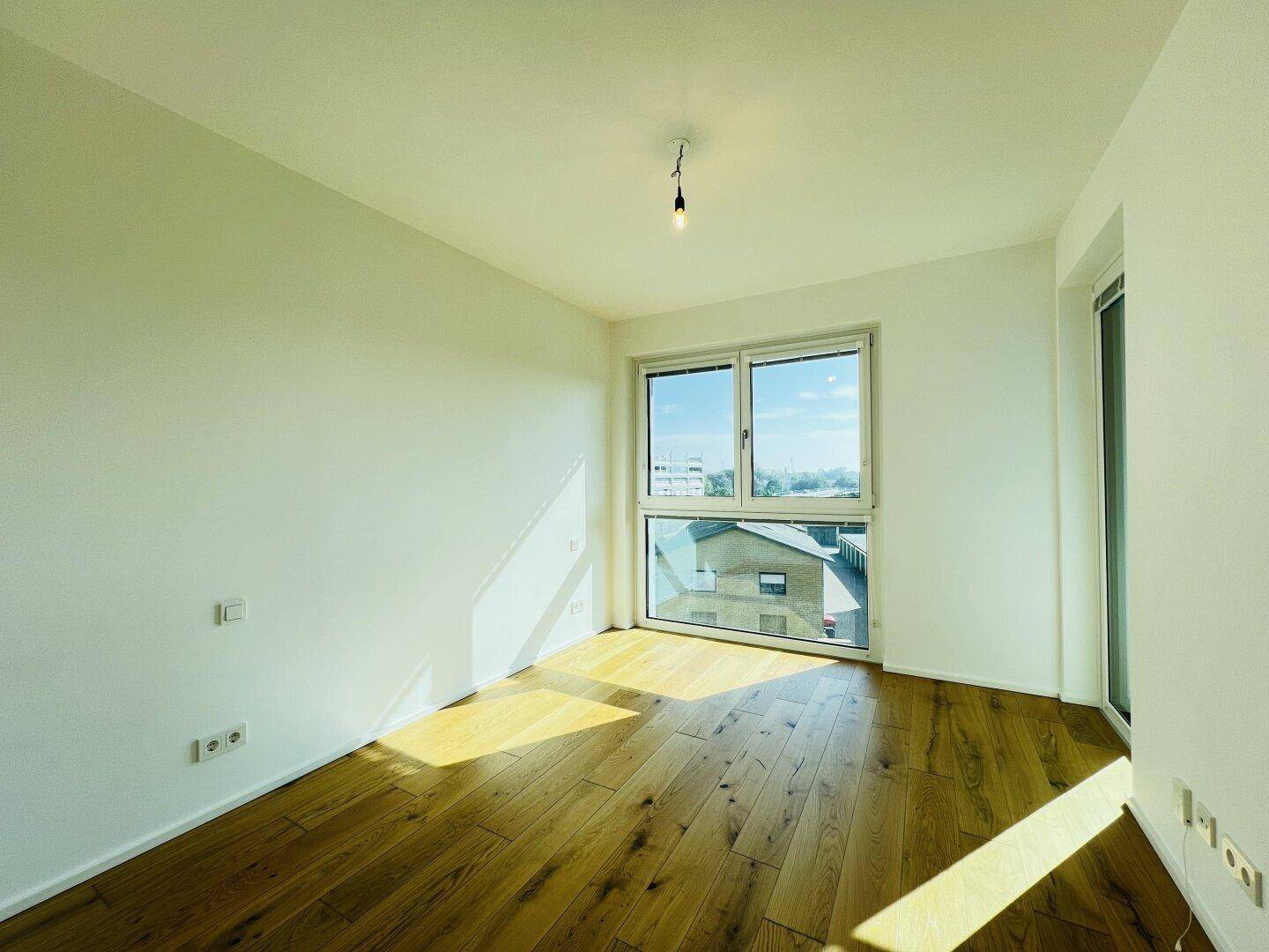 Helles, leeres Zimmer mit Holzboden und großem Fenster, das viel Tageslicht hereinlässt.