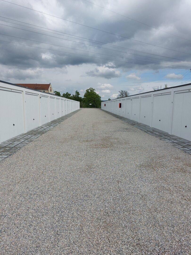 Lange Reihe von geschlossenen Garagenboxen mit weißen Toren entlang eines Schotterwegs unter bewölktem Himmel.