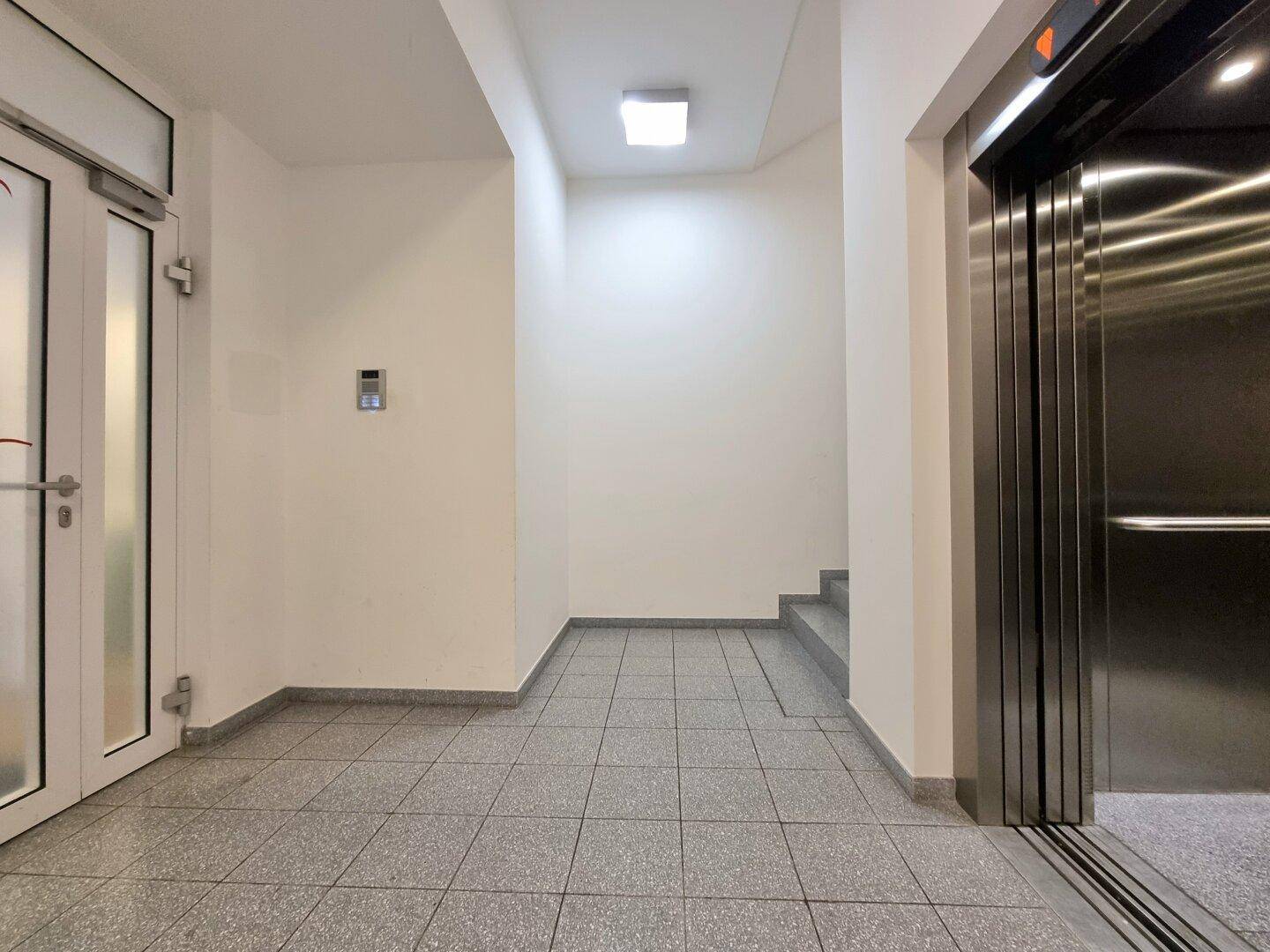 Heller Flur mit Fliesenboden, Aufzug und Treppenaufgang in einem Mehrfamilienhaus.