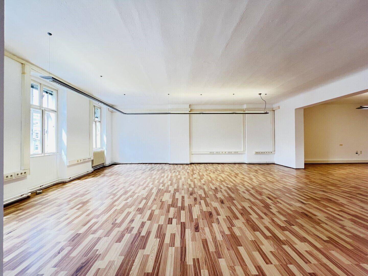 Geräumiger, leerer Raum mit hellem Laminatboden und großen Fenstern, ideal für verschiedene Nutzungen.
